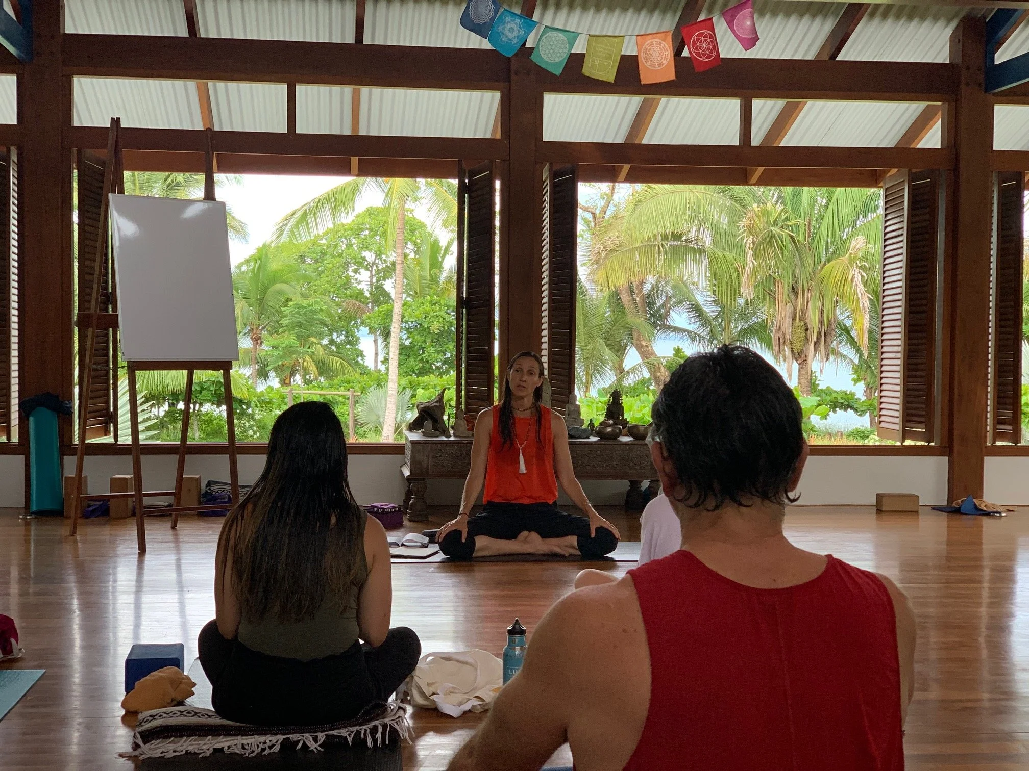 A woman in orange and black clothing is sitting cross-legged on a wooden floor during a yoga or meditation class, with her eyes closed. Several other people are seated on the floor around her, facing her, in a spacious room with open windows and lush