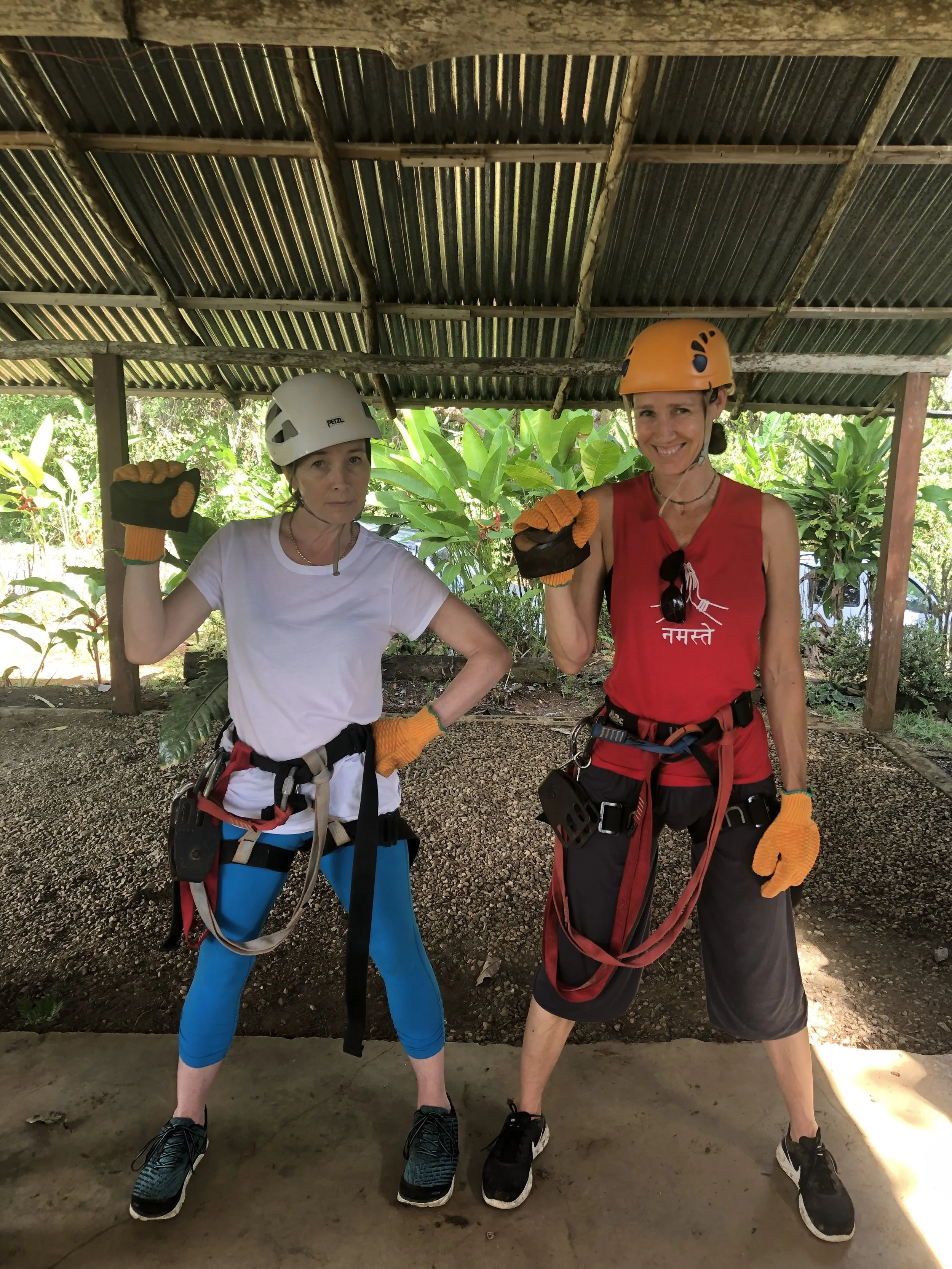 Two women dressed in climbing gear, standing under a roofed outdoor structure with greenery in the background, smiling and showing their fists in a friendly pose.