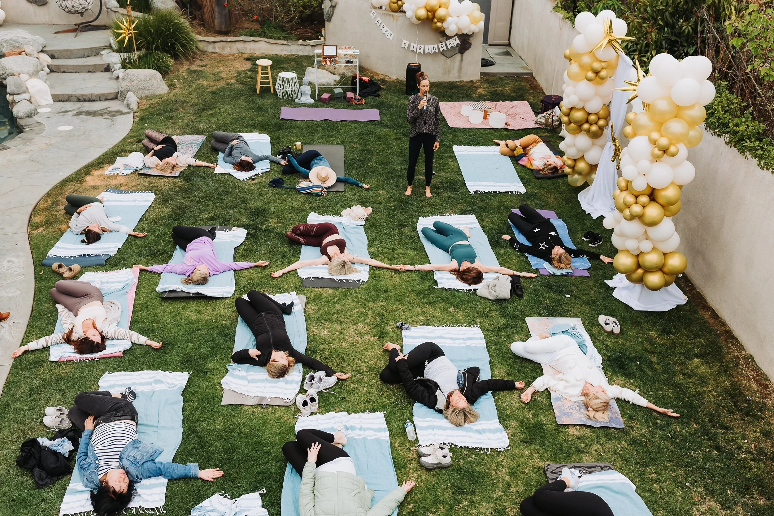 People participating in an outdoor yoga or meditation session led by Vivica Schwartz on a grassy backyard, lying on mats. Private birthday event.