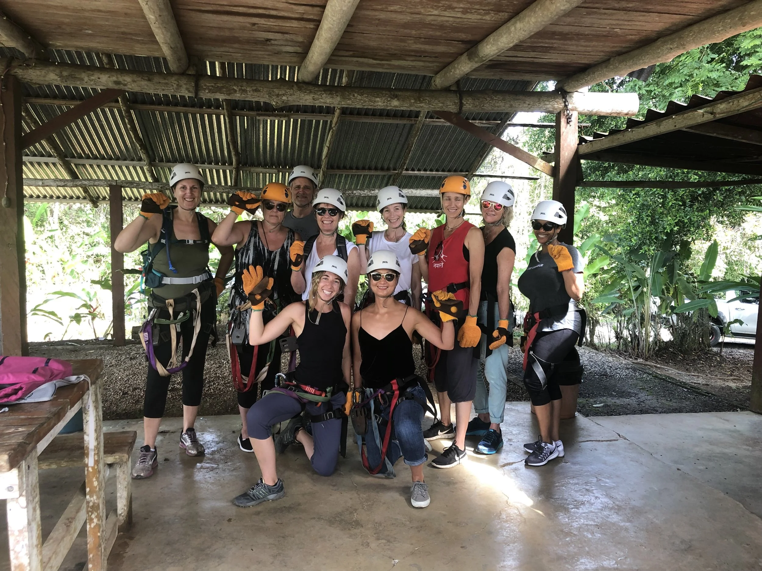 A group of people wearing helmets and gloves, standing together and smiling, in an outdoor shelter with a corrugated roof, surrounded by greenery.