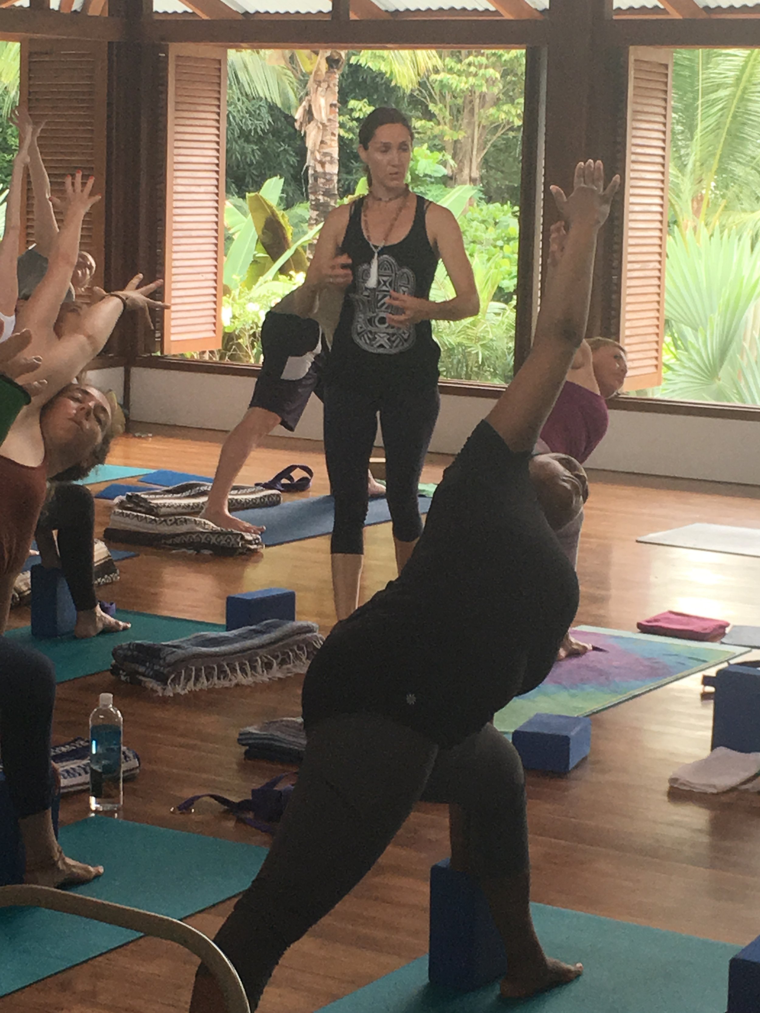 A group of people participating in a yoga class in a room with open windows showing lush greenery outside. The instructor is standing and talking.  Yoga retreat in Costa Rica led by Vivica Schwartz. 