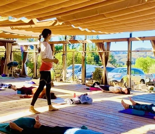 Vivica Schwartz teaching a yoga class taking place on a wooden deck with several participants and a scenic outdoor background with trees and parked cars.