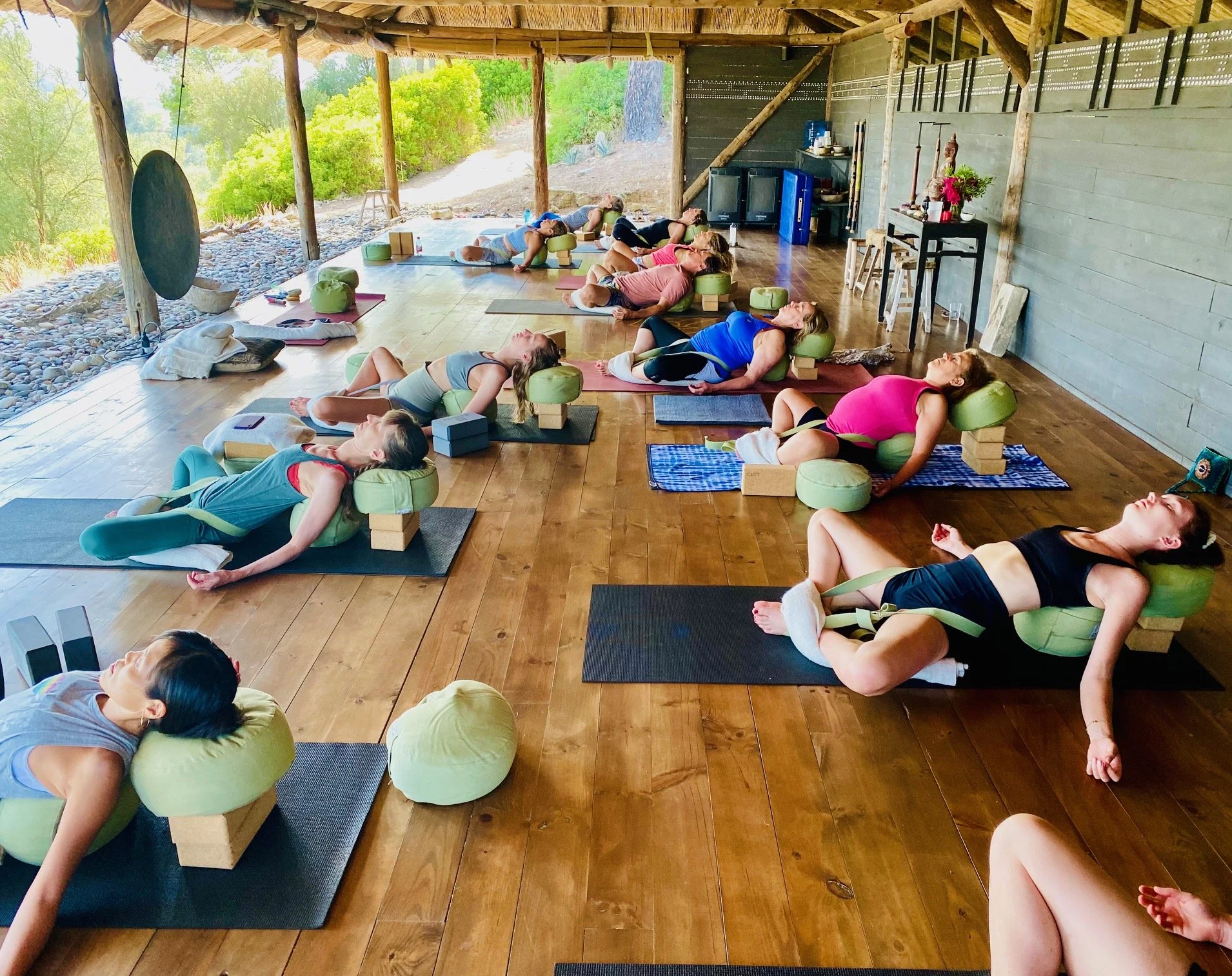 Group of people at a Yoga retreat in Mallorca led by Vivica Schwartz, practicing yoga in a rustic outdoor sheltered space, lying on yoga mats with bolsters and blocks, surrounded by natural scenery including rocks and trees.