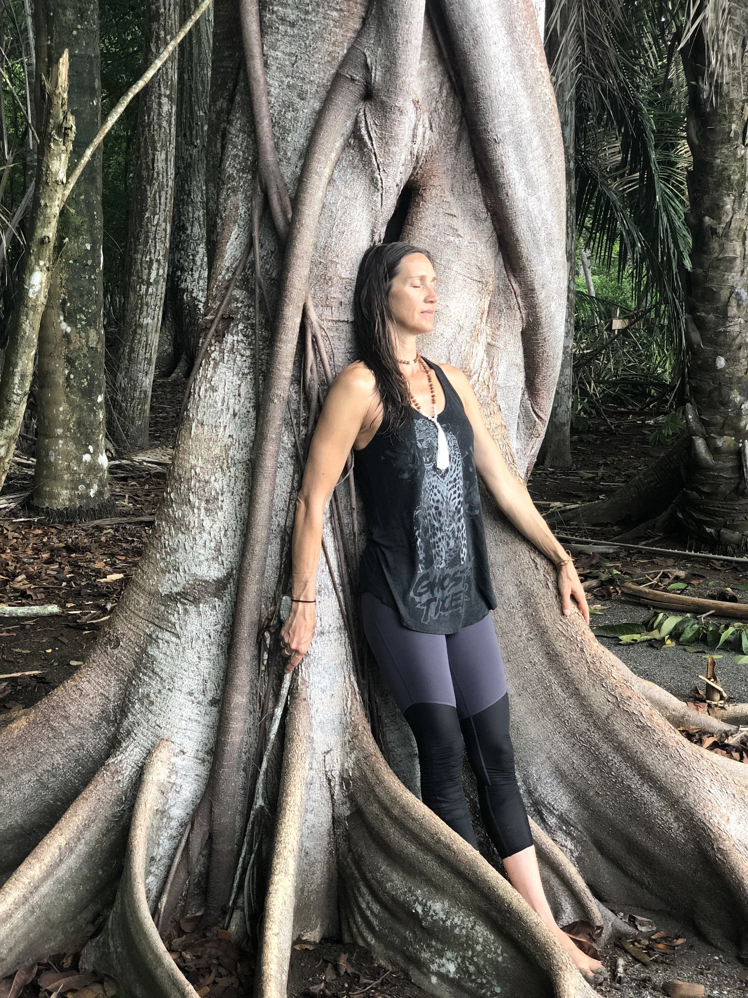 A woman with long dark hair, wearing a black sleeveless top with leopard print and black leggings, is leaning against a large tree with exposed roots in a dense jungle. Yoga retreat in Costa Rica led by Vivica Schwartz. 