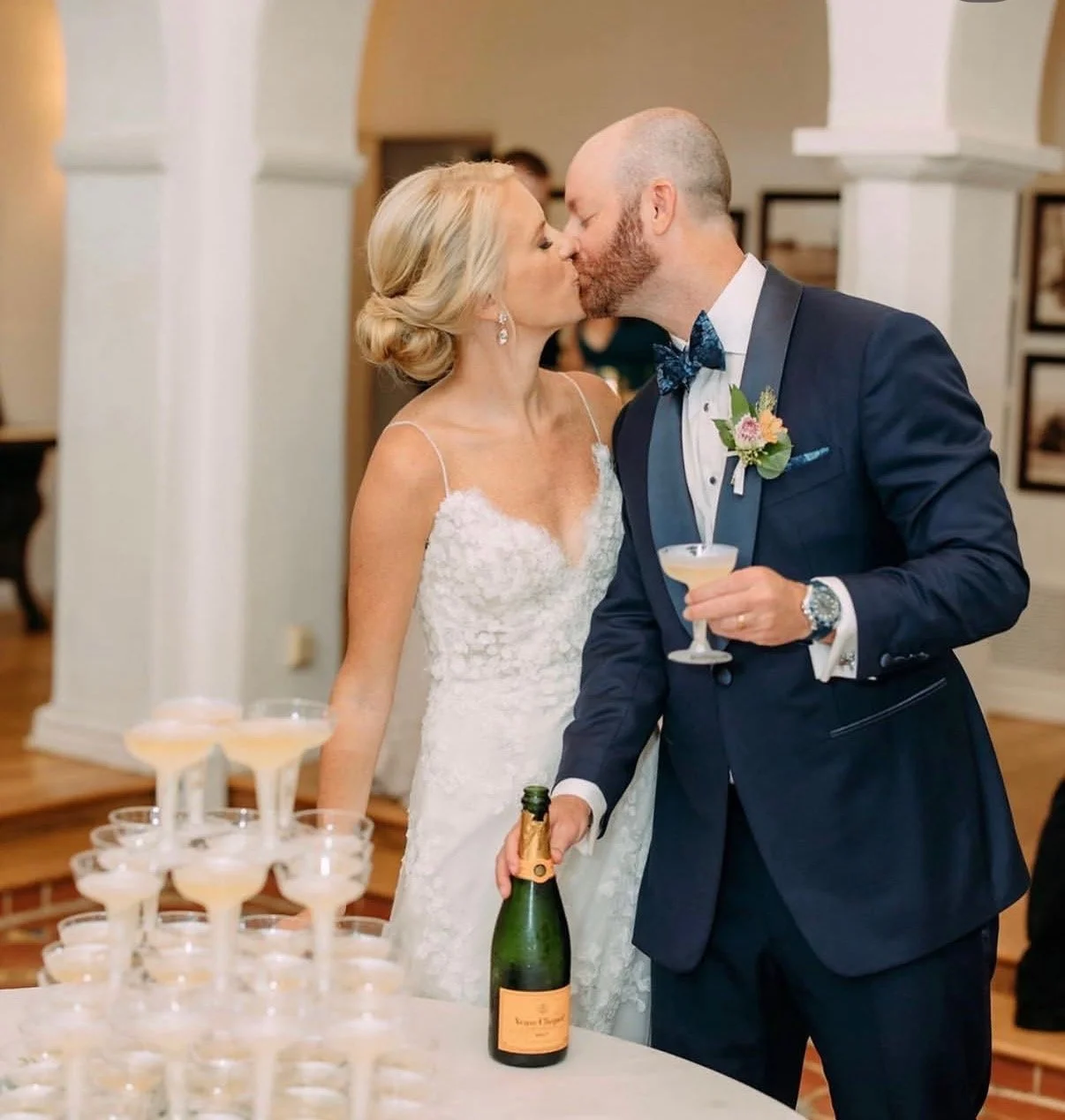 A bride and groom sharing a kiss at their wedding reception, standing next to a table with champagne glasses and a bottle of champagne.