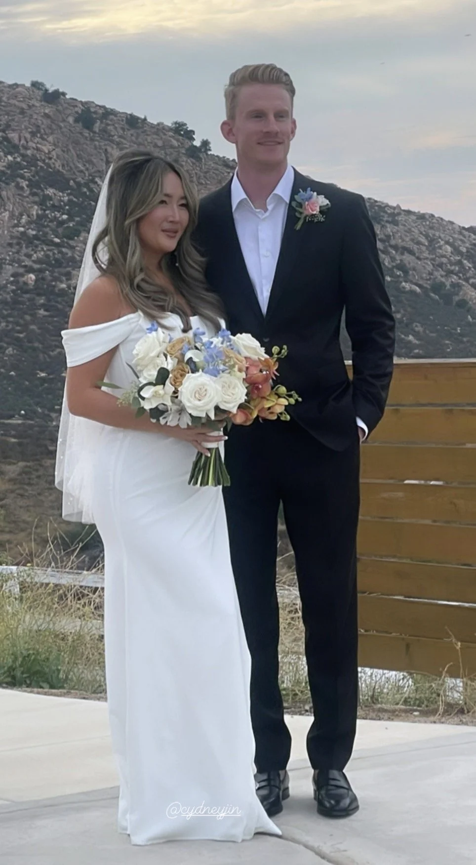 A bride and groom posing outdoors on their wedding day, with rocky hills and a cloudy sky in the background. The bride wears a white off-shoulder wedding dress and holds a bouquet of flowers, while the groom is dressed in a black suit with a white sh
