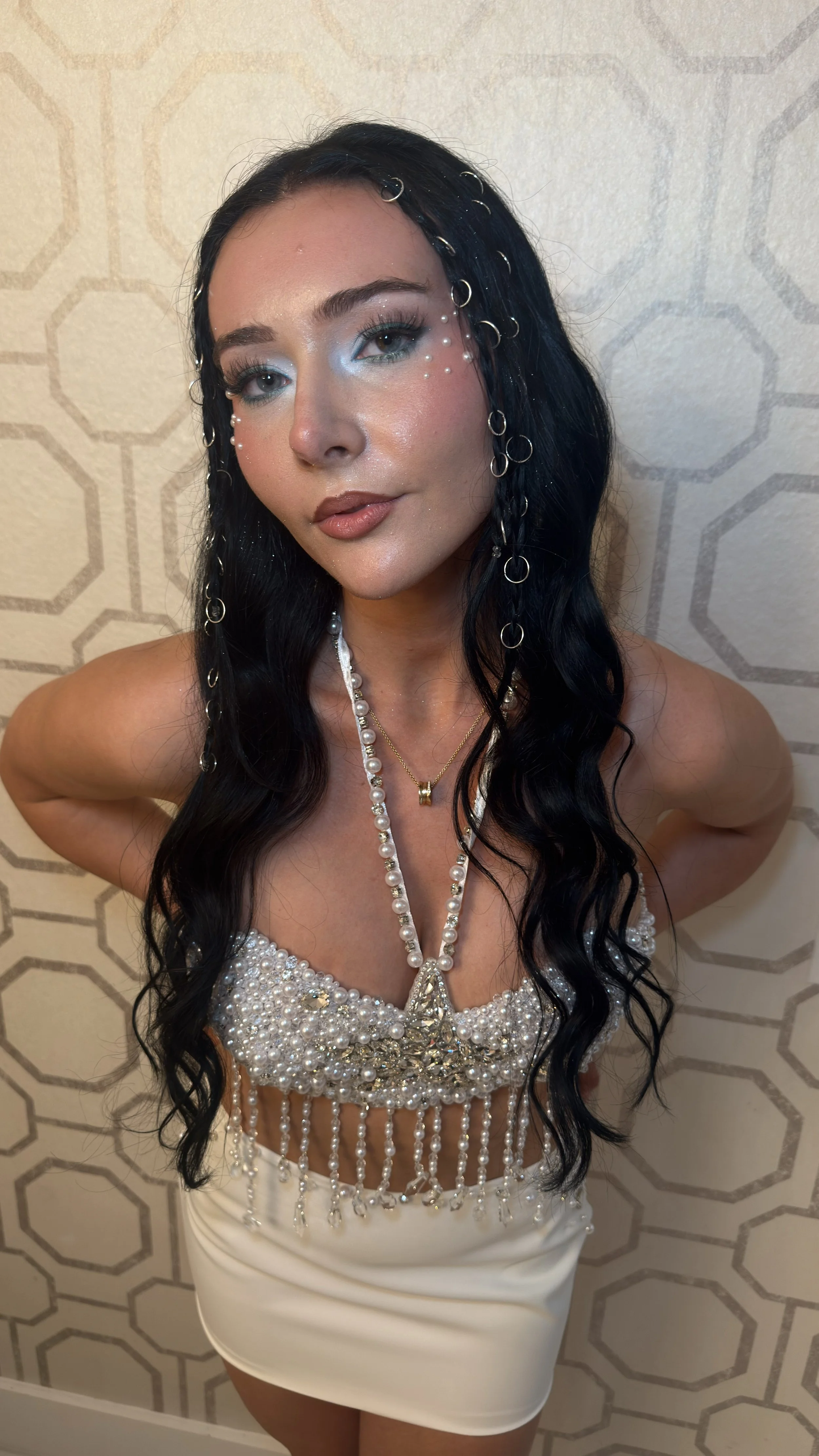 A woman with long wavy black hair and blue eyeshadow, wearing pearl and silver jewelry, a pearl embellished top, and a white skirt, standing in front of a patterned wall.