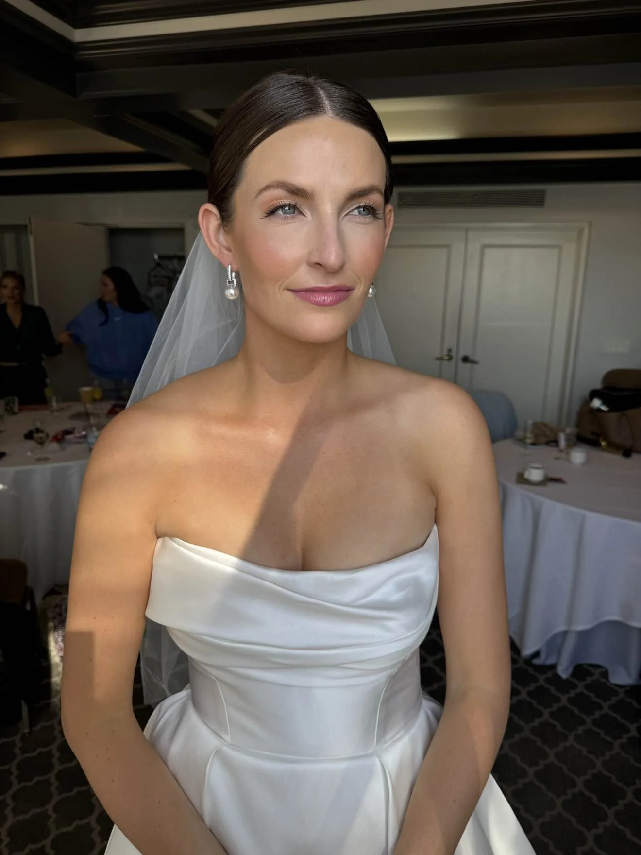 Close-up of a woman in a wedding dress with a veil, earrings, and makeup, smiling slightly, in a room with a table and people in the background.