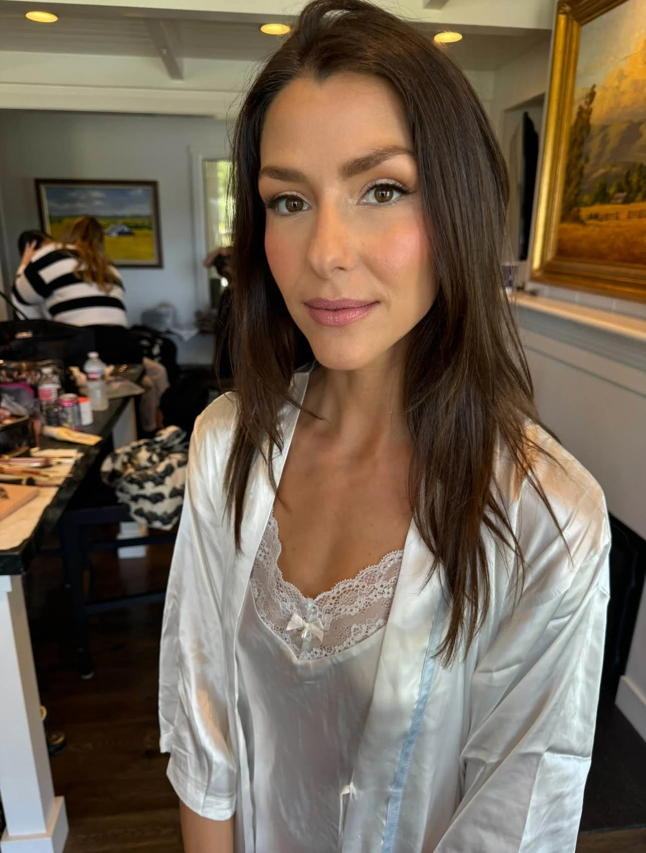 Woman with long dark hair wearing a satin robe with lace trim, preparing for her makeup or hair in a room with painting on the wall and makeup supplies on a table.