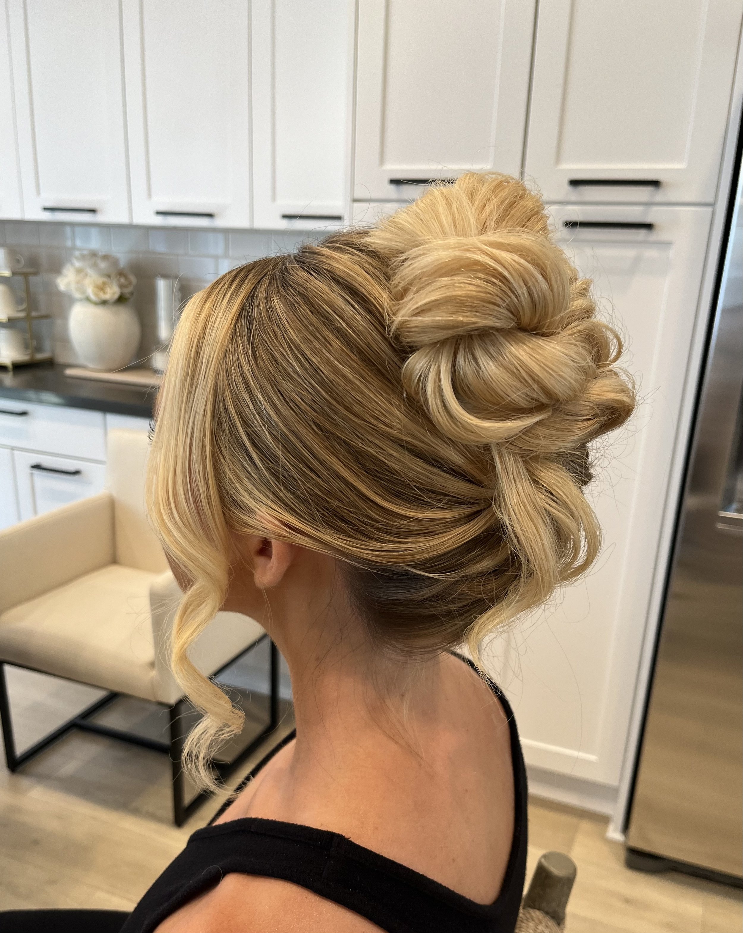 A woman with blonde hair styled in an elegant updo featuring soft curls at the ends, set indoors in a modern space with white cabinetry and a vase of white flowers in the background.