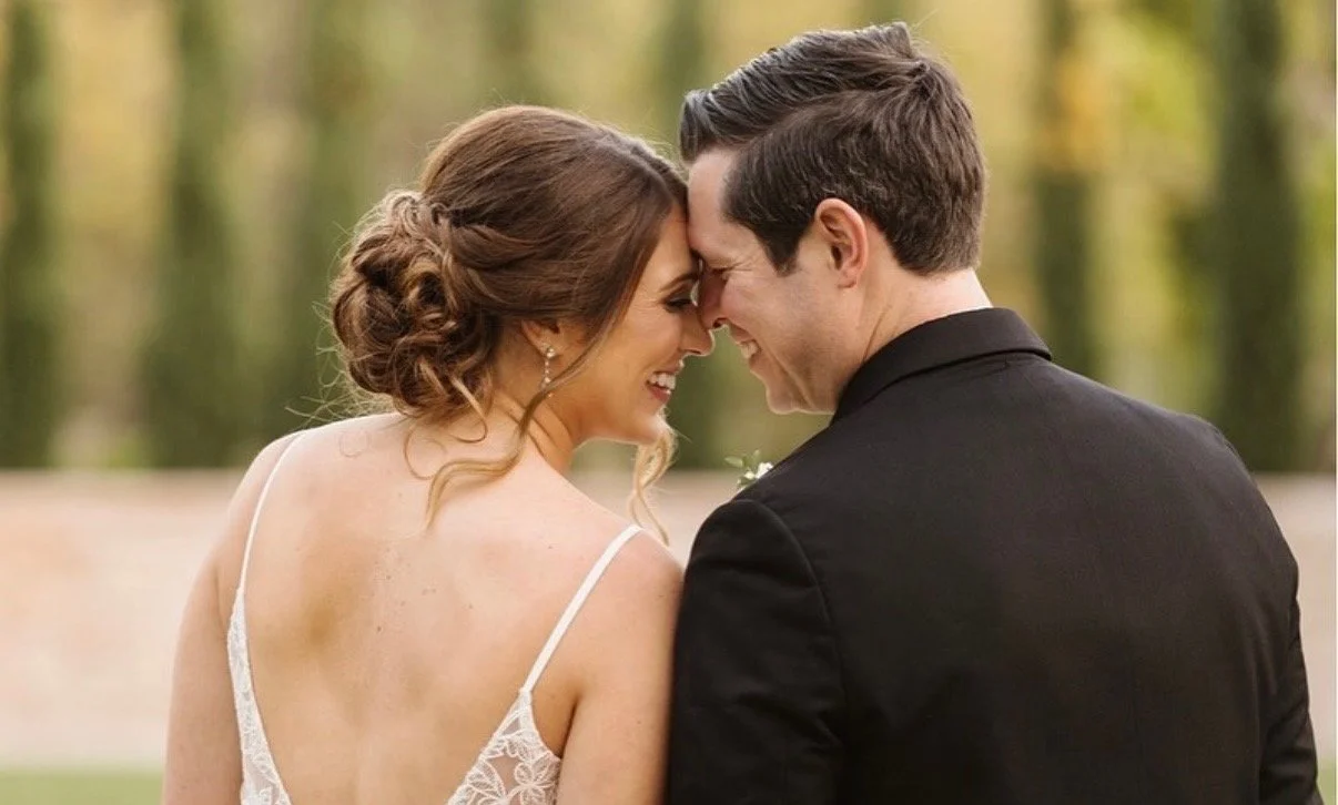 A bride and groom with foreheads touching and smiling outdoors during a wedding.