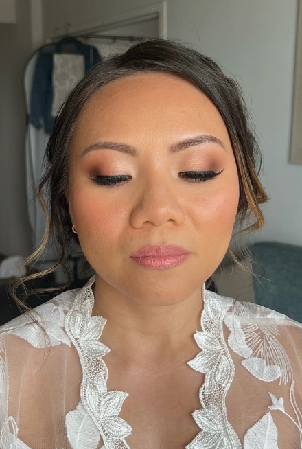Close-up of a woman with makeup, earrings, and a sheer lace top, looking down with a neutral expression in a room with a mirror and curtains in the background.