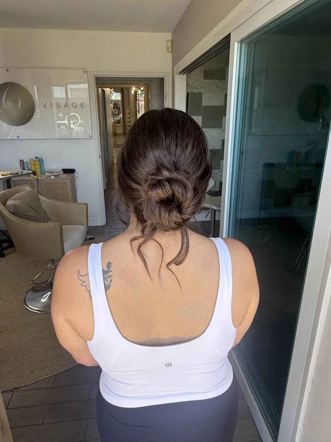 A woman with dark brown hair styled in a loose bun, wearing a white tank top, standing indoors in front of a glass door, with a tattoo on her shoulder.