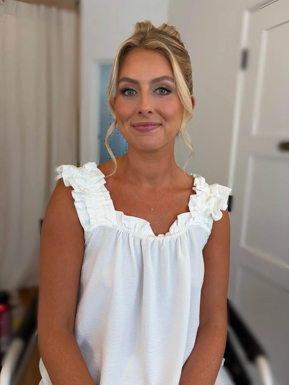 A woman with blonde hair styled in an updo with loose curls, wearing a white dress with ruffled straps and a small cross necklace, standing indoors with a blurred background of white walls and a closed door.