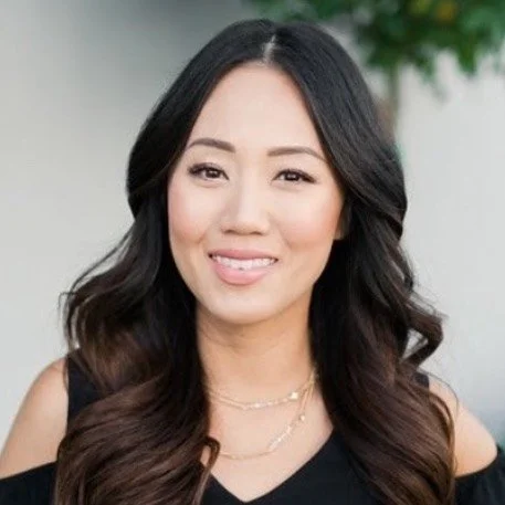A woman with long dark hair smiling, wearing a black top and layered necklaces.