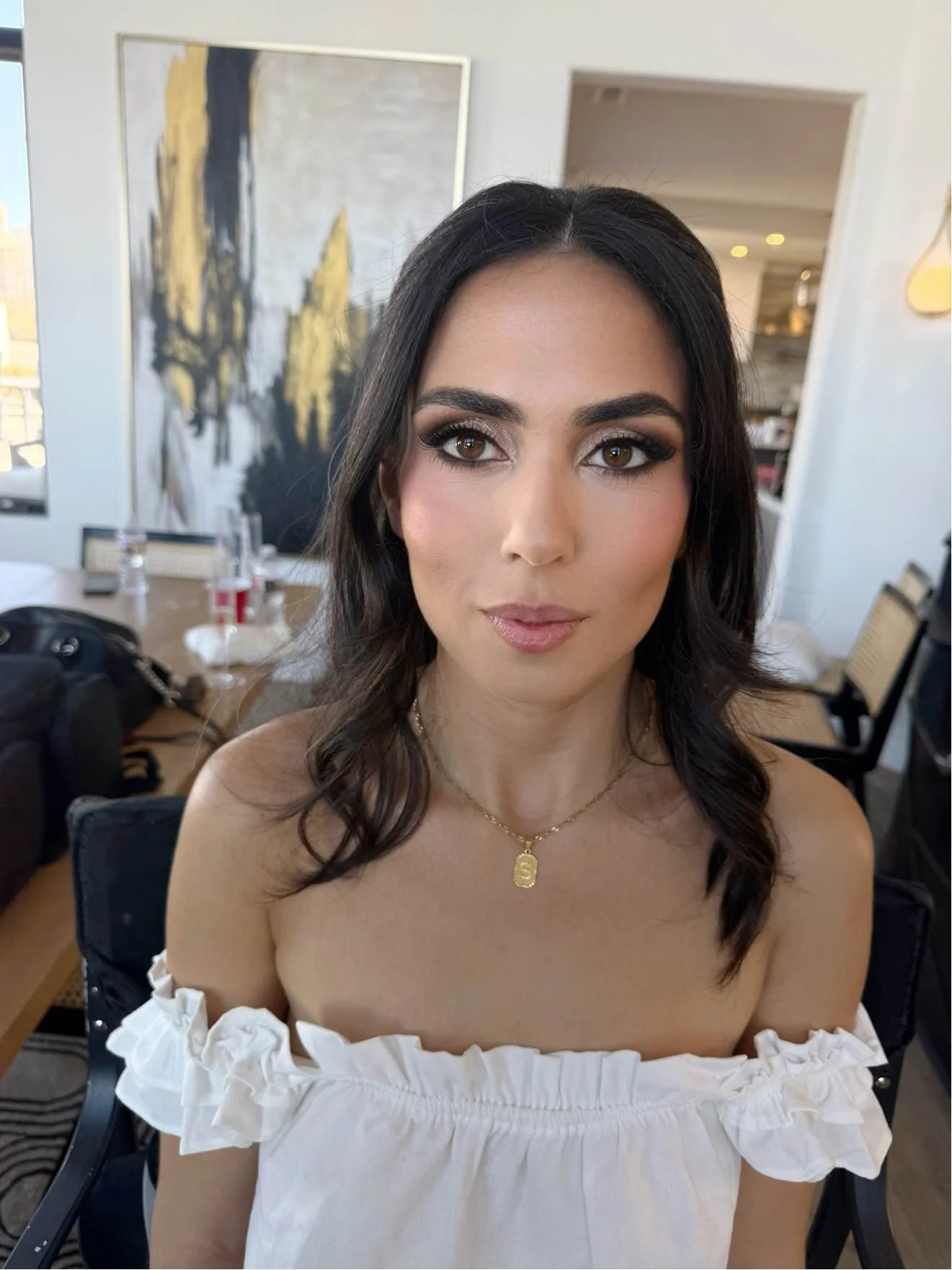 Close-up of a woman with dark brown hair, brown eyes, and makeup, wearing a white off-shoulder top with ruffled sleeves and a gold necklace with a letter 'S' pendant, sitting in an indoor setting with artwork on the wall.