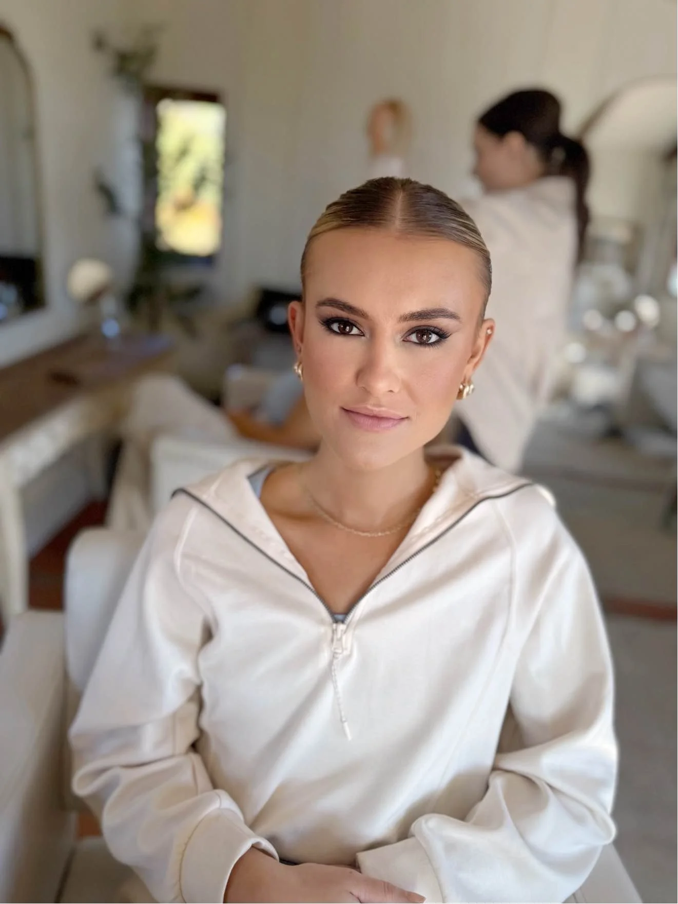 A young woman with blonde hair and makeup, wearing a white zip-up jacket, sitting indoors with a blurred background of another woman with dark hair and a mirror.