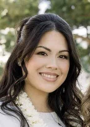 Close-up of a young woman with long dark curly hair smiling outdoors with greenery in the background.