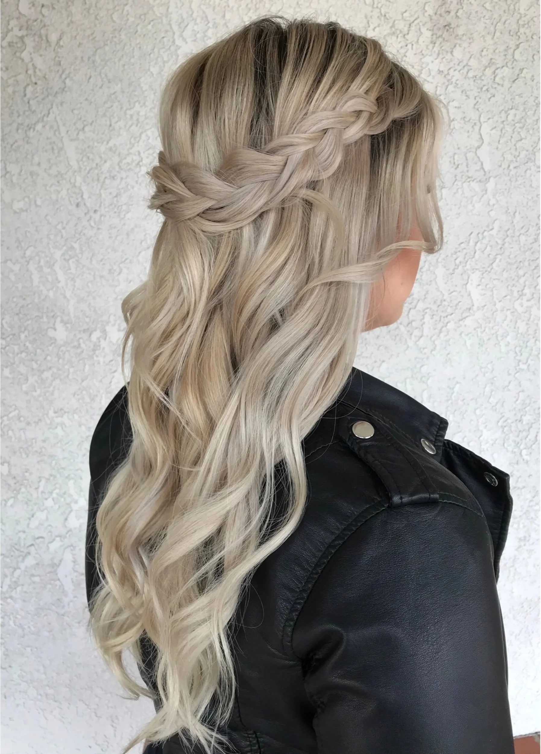 Blonde woman with loose and braided hair wearing a black leather jacket, photographed against a textured wall.