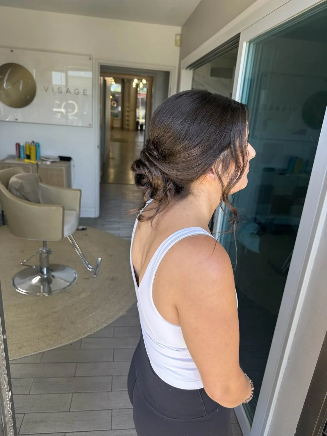 A woman with brown hair styled in a low, twisted bun looks at a glass door inside a modern salon or spa.