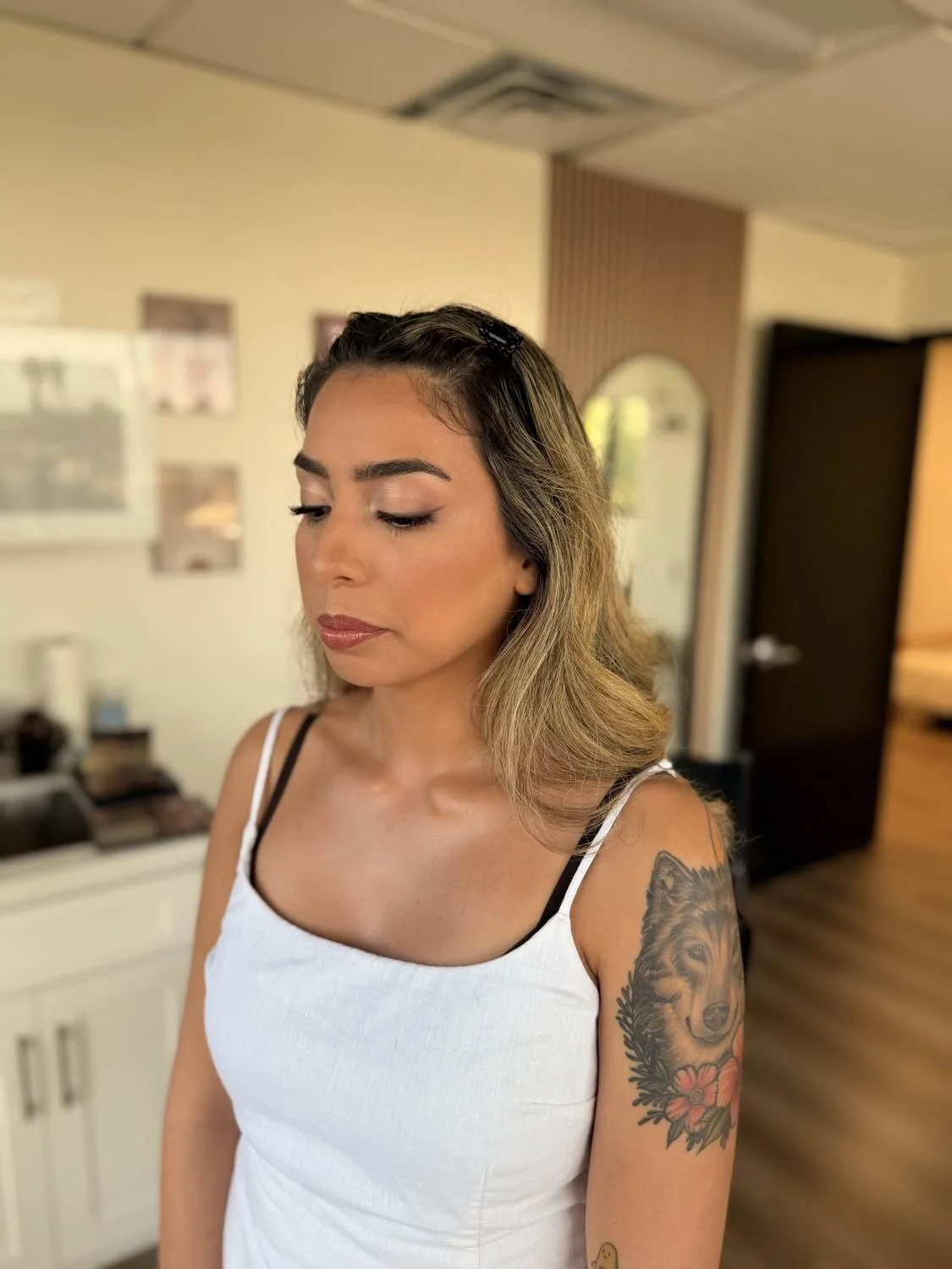 A woman with shoulder-length wavy hair, wearing a white tank top with black straps, looking down with closed eyes, showing a detailed tattoo of a wolf on her upper arm with flowers.