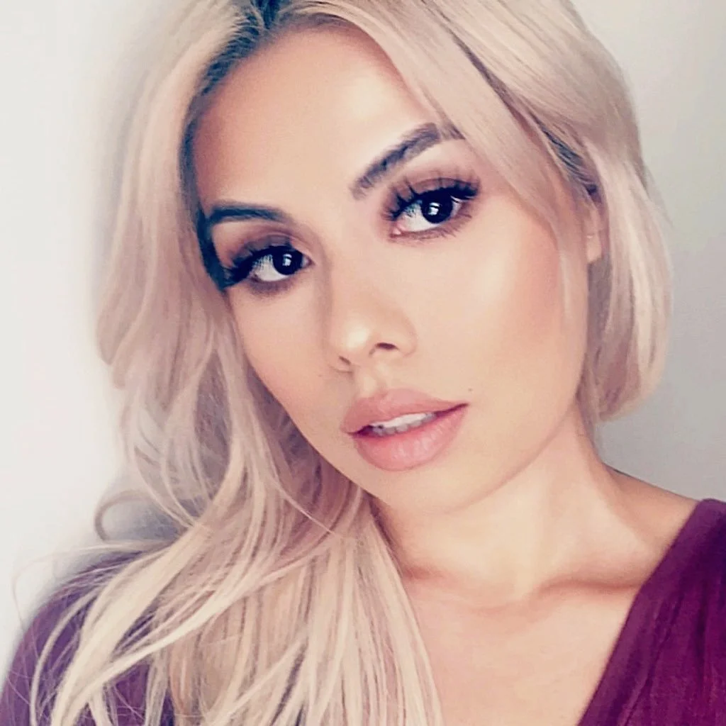 Close-up of a woman with platinum blonde hair, makeup with bold eyeliner and nude lipstick, wearing a purple top.