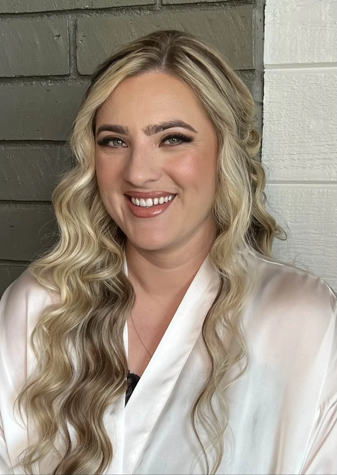 A woman with long, wavy blonde hair smiling showing her teeth, wearing a light-colored satin robe, standing in front of a background of gray and white wooden panels.
