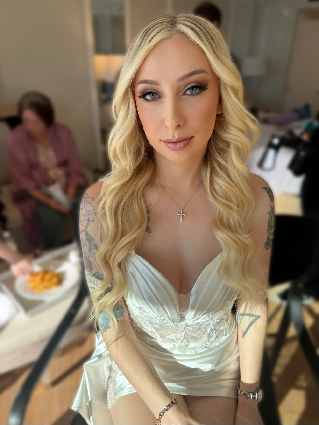 A woman with long blonde hair and tattoos wearing a white dress and a cross necklace sitting in a room, with a blurred background of people and furniture.