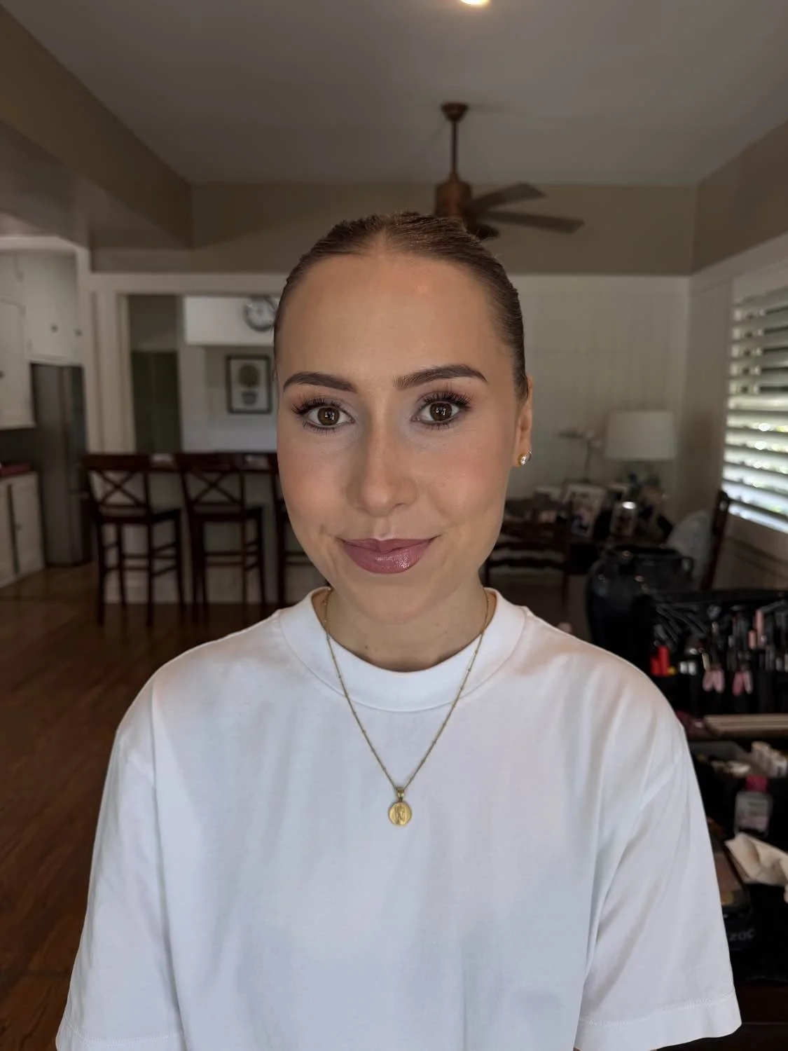 Close-up of a smiling woman with brown hair pulled back, wearing a white shirt and gold jewelry, in a well-lit living room with wooden floors and kitchen area in the background.