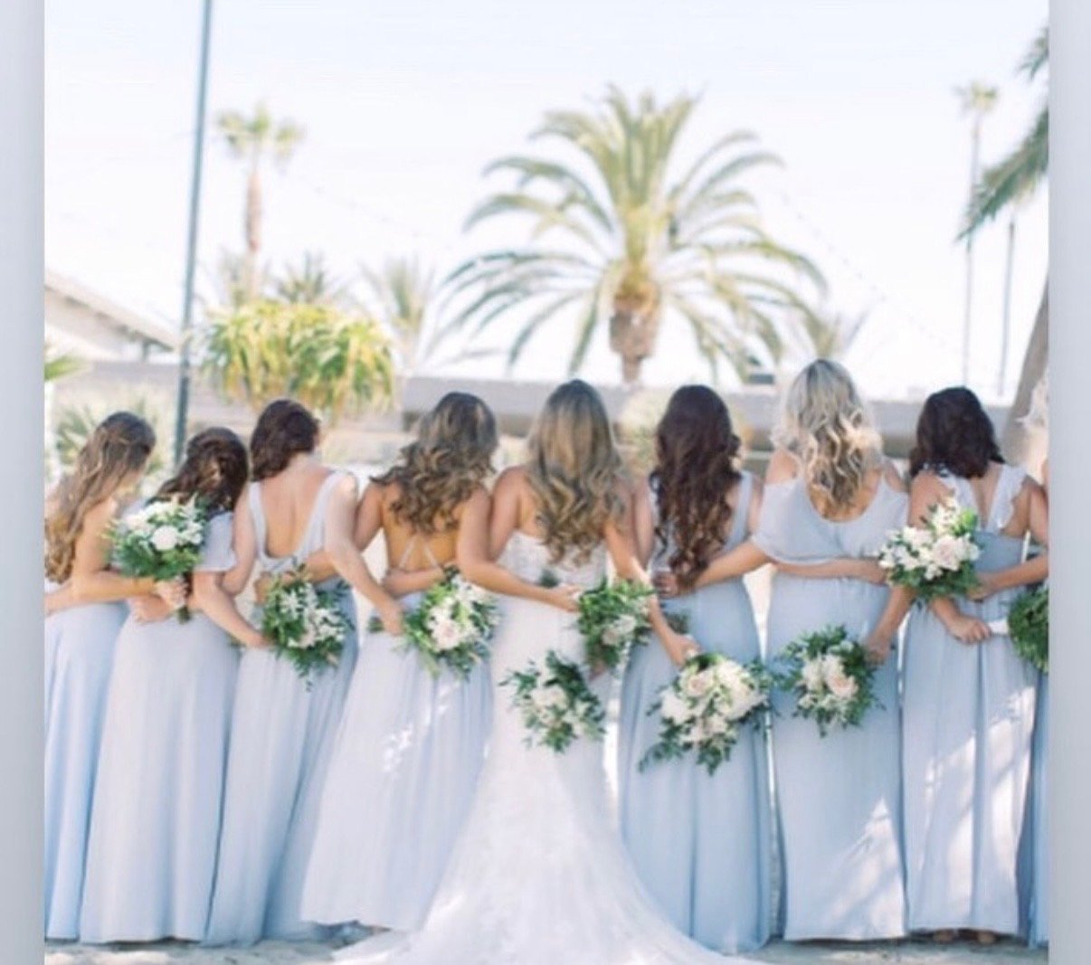 Line of women in light blue dresses holding bouquets, standing arm-in-arm outdoors with palm trees in background, suggesting a wedding or special event.