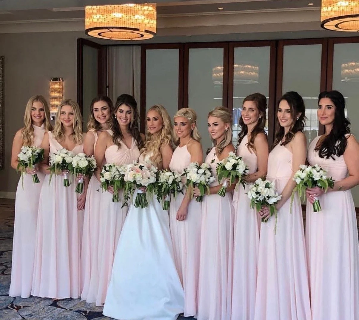 A group of eleven women in light pink dresses holding bouquets of white flowers posing indoors.