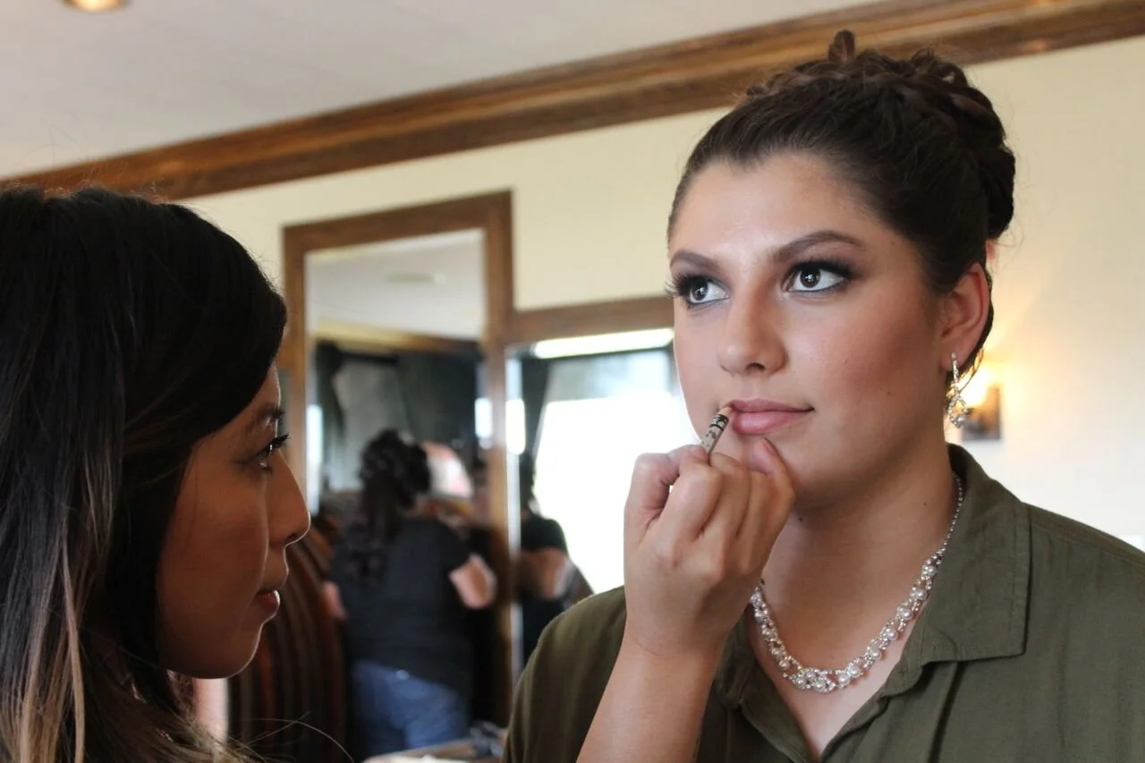 A makeup artist applying lip makeup to a young woman with styled hair and jewelry in a softly lit room with other people in the background.