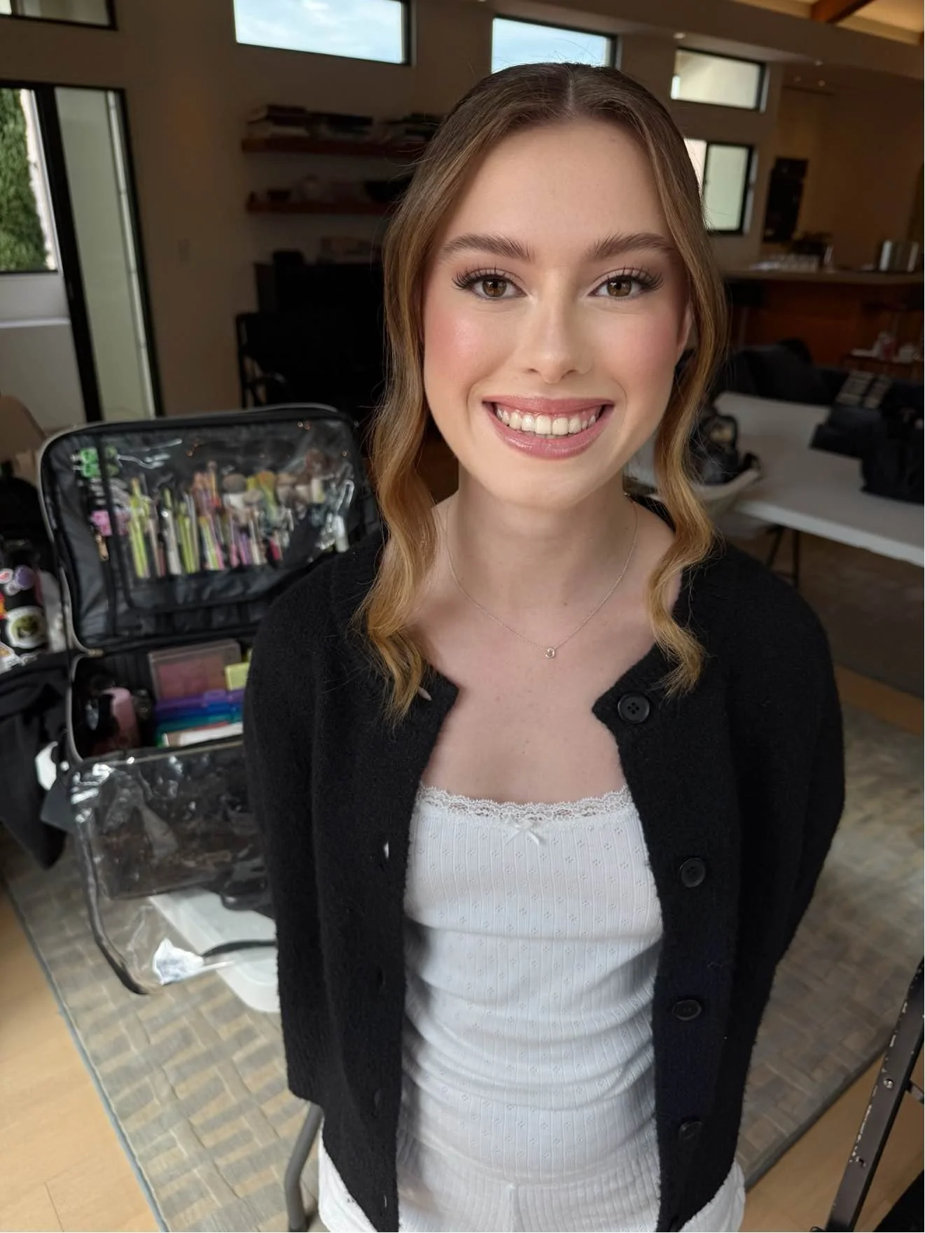 A young woman with light skin and brown wavy hair styled in loose curls is smiling at the camera. She is wearing a white top with lace and a black buttoned cardigan. The background shows a living space with shelves, windows, and makeup tools and supp