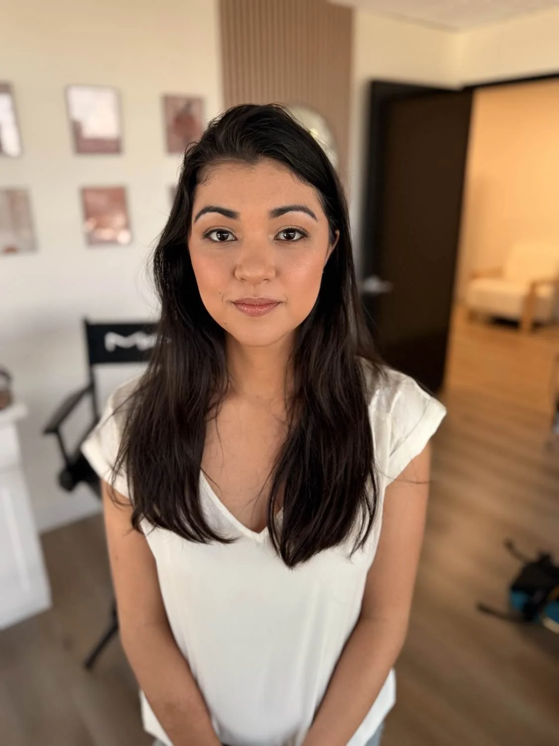 A young woman with long dark hair in a white top standing indoors with a blurred background including wall art and furniture.