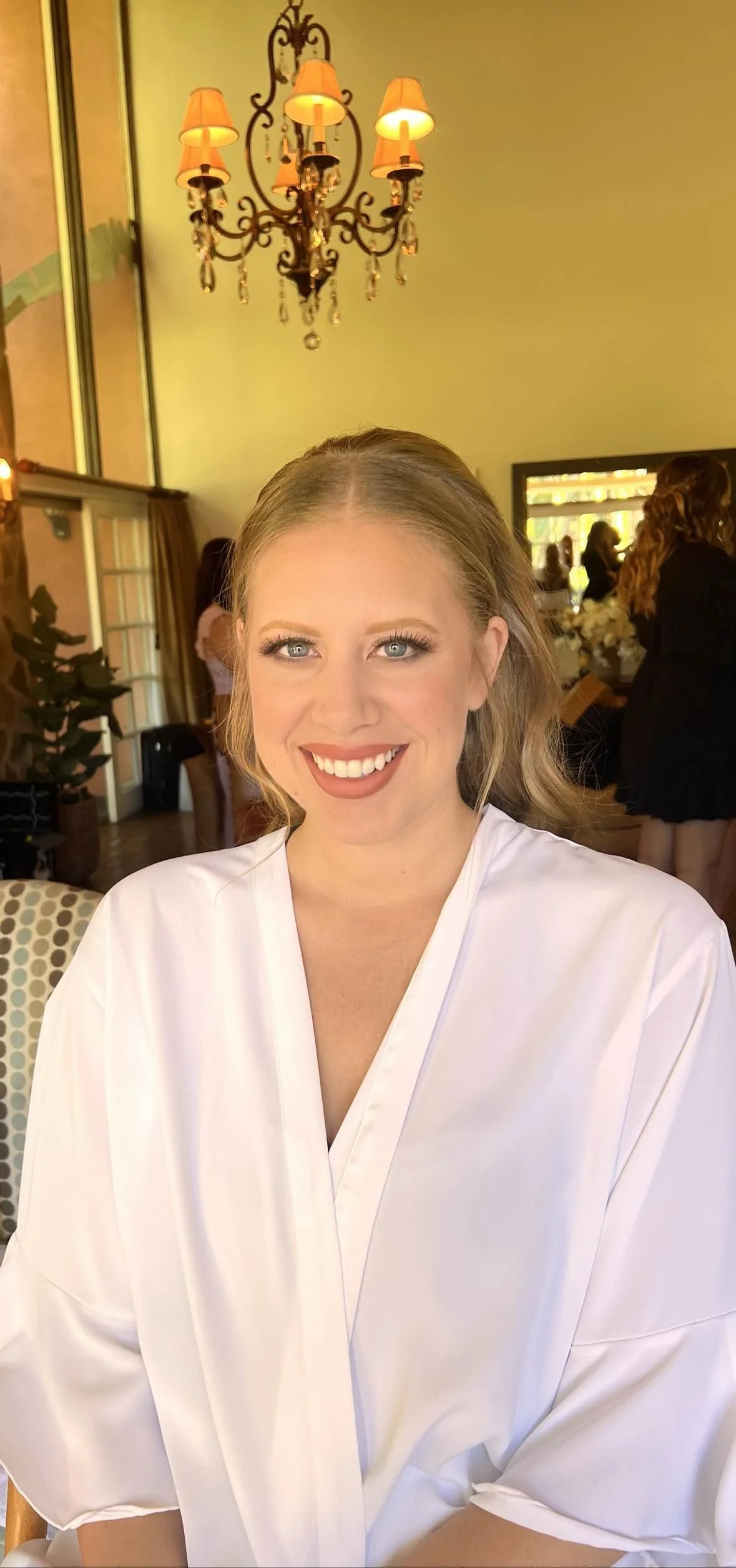 A woman with blonde hair, blue eyes, and a wide smile, wearing a white robe, in a room with yellow walls, a chandelier, and other people in the background.