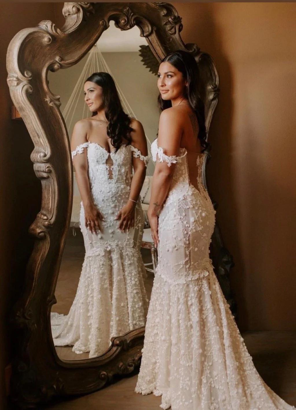 A woman in a lace wedding dress stands looking at herReflection in an ornate wooden mirror.
