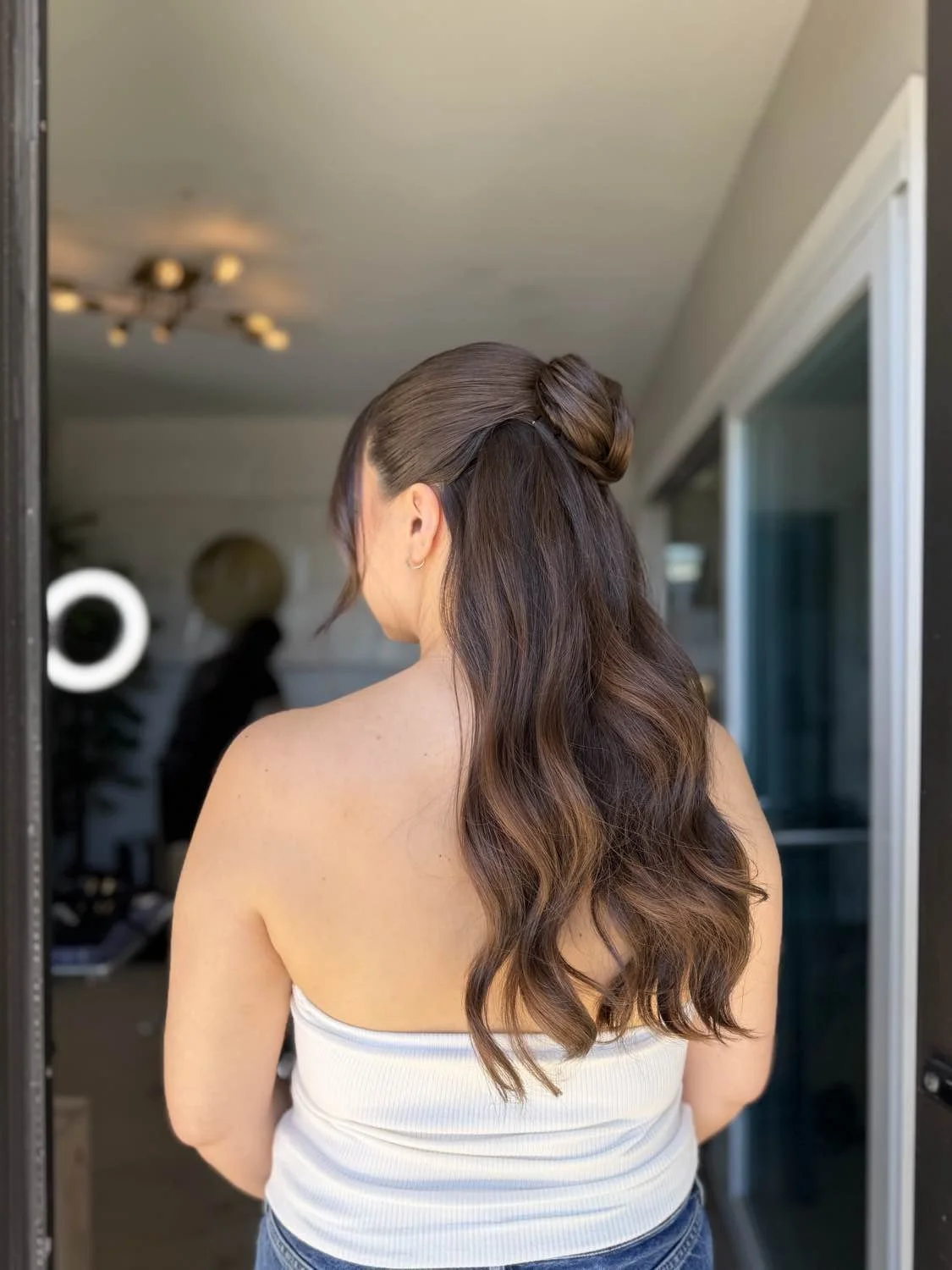 Back view of a woman with half-up hairstyle, wearing a white tank top, standing in a doorway with a room and window in the background.