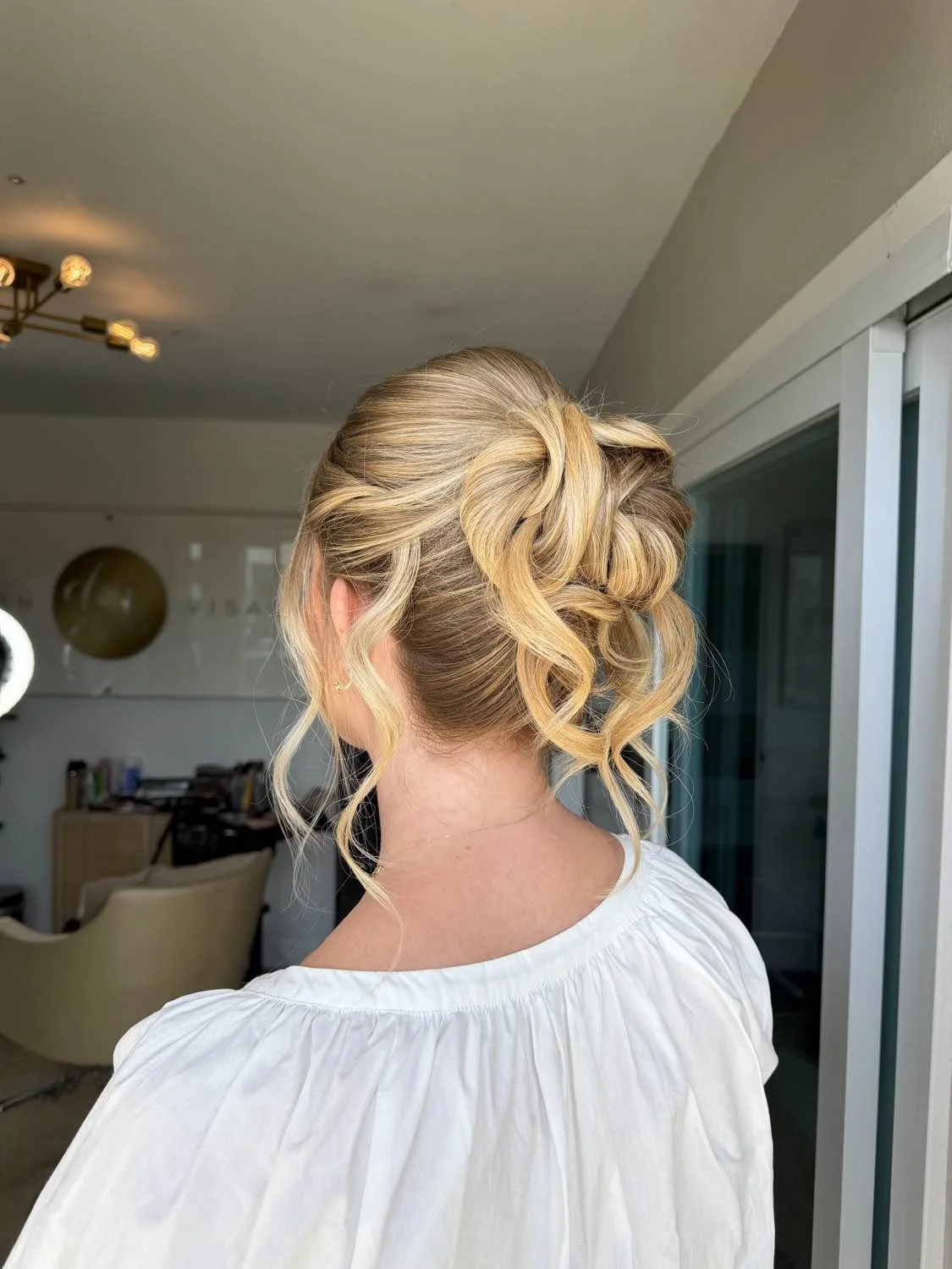 Woman with blonde hair styled in an elegant updo with loose, curled strands, seen from the back in a well-lit room.