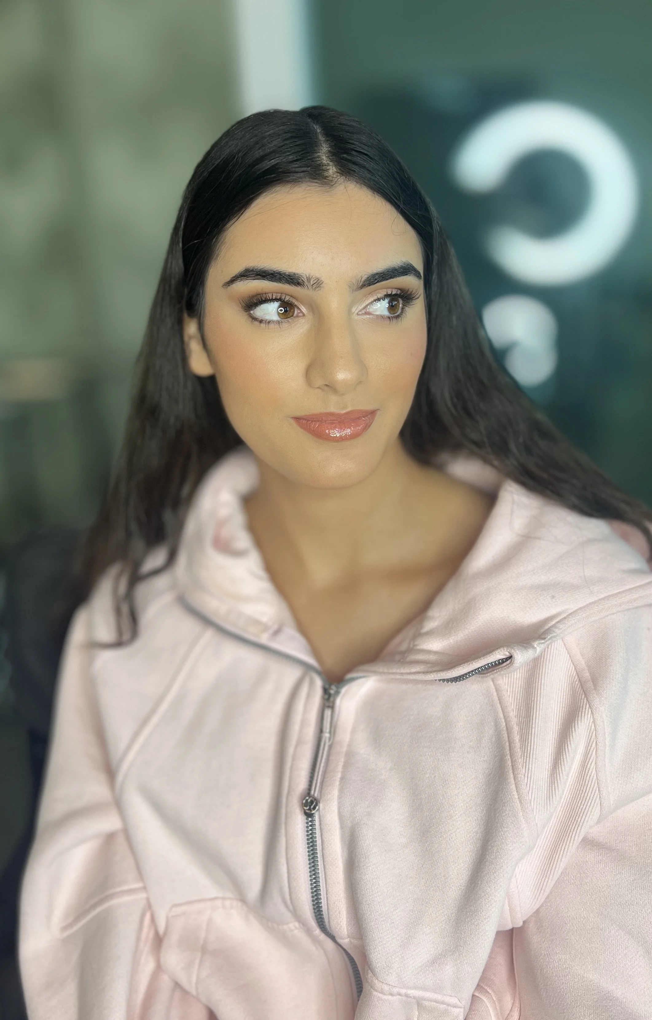 Close-up of a woman with long dark hair, brown eyes, and makeup, wearing a pink zip-up hoodie.