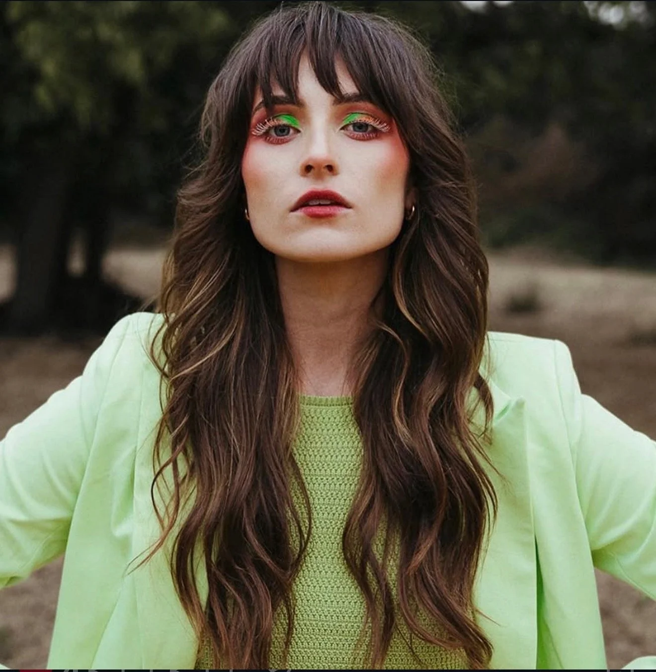 A woman with long wavy brown hair and bangs wearing neon green makeup and a matching green blazer outdoors with trees in the background.