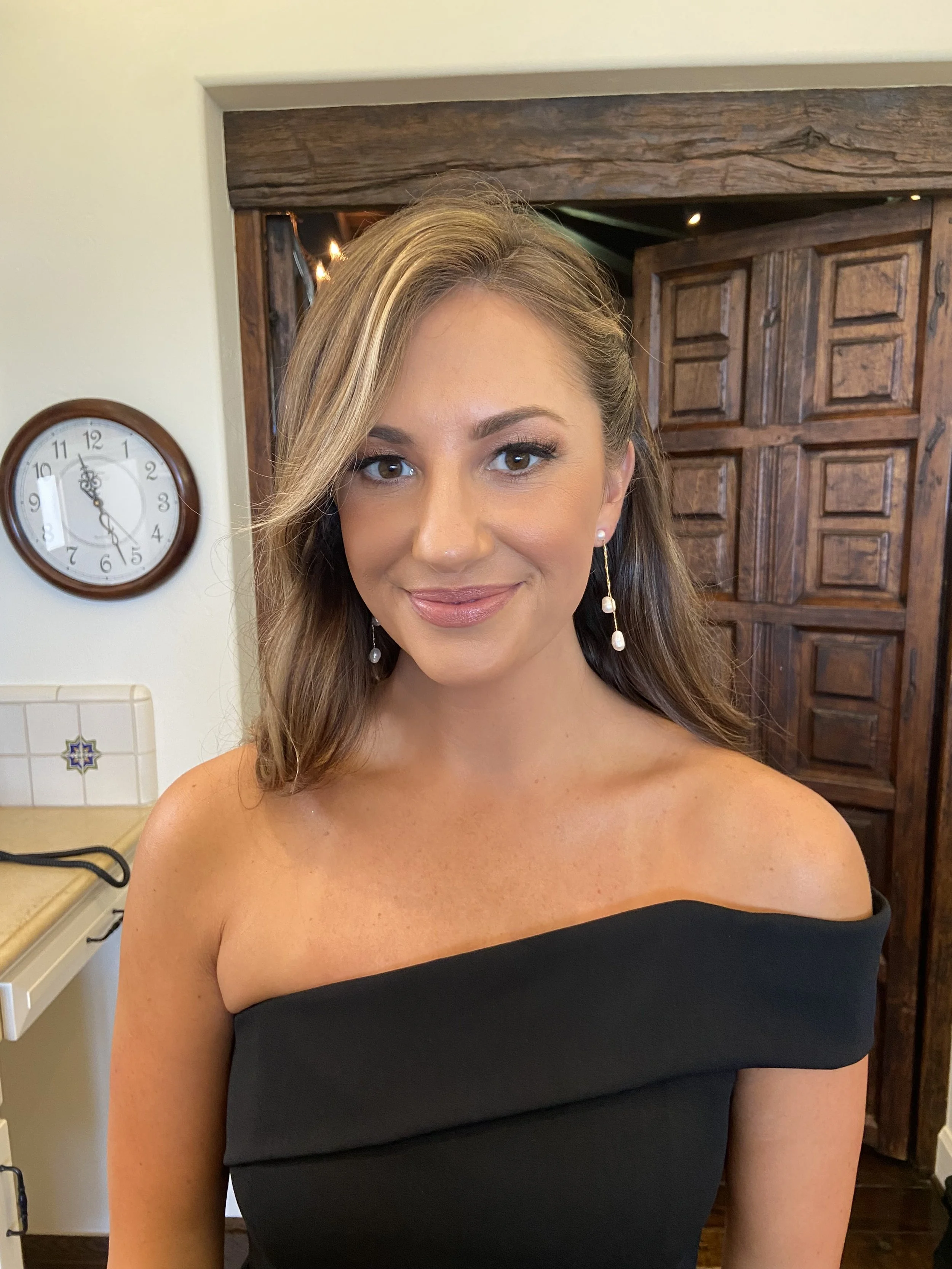 Portrait of a woman with long brown hair in a black off-shoulder top, smiling in a room with a wooden door and a clock on the wall behind her.