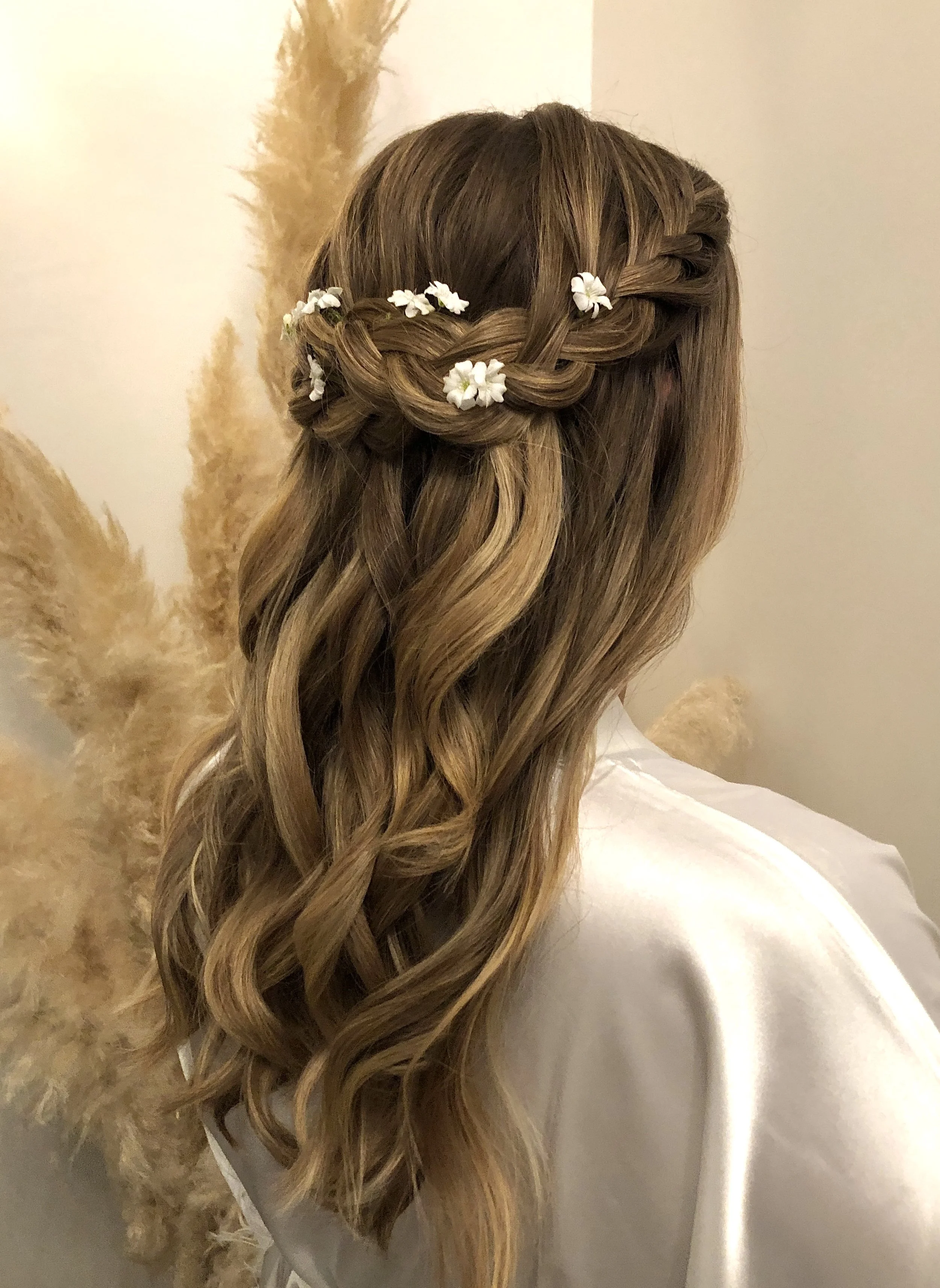 Close-up of a woman with long, wavy blonde hair styled in a braided hairstyle with small white flowers woven into the braid.