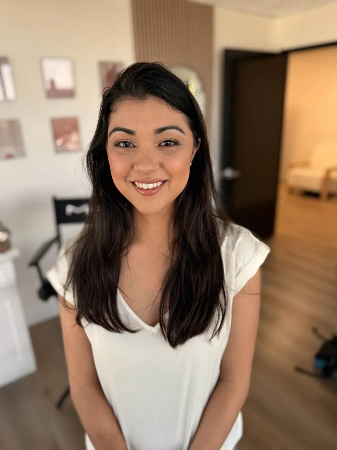 A young woman with dark hair smiling, wearing a white top, standing indoors with a blurred background of wall art, furniture, and room decor.