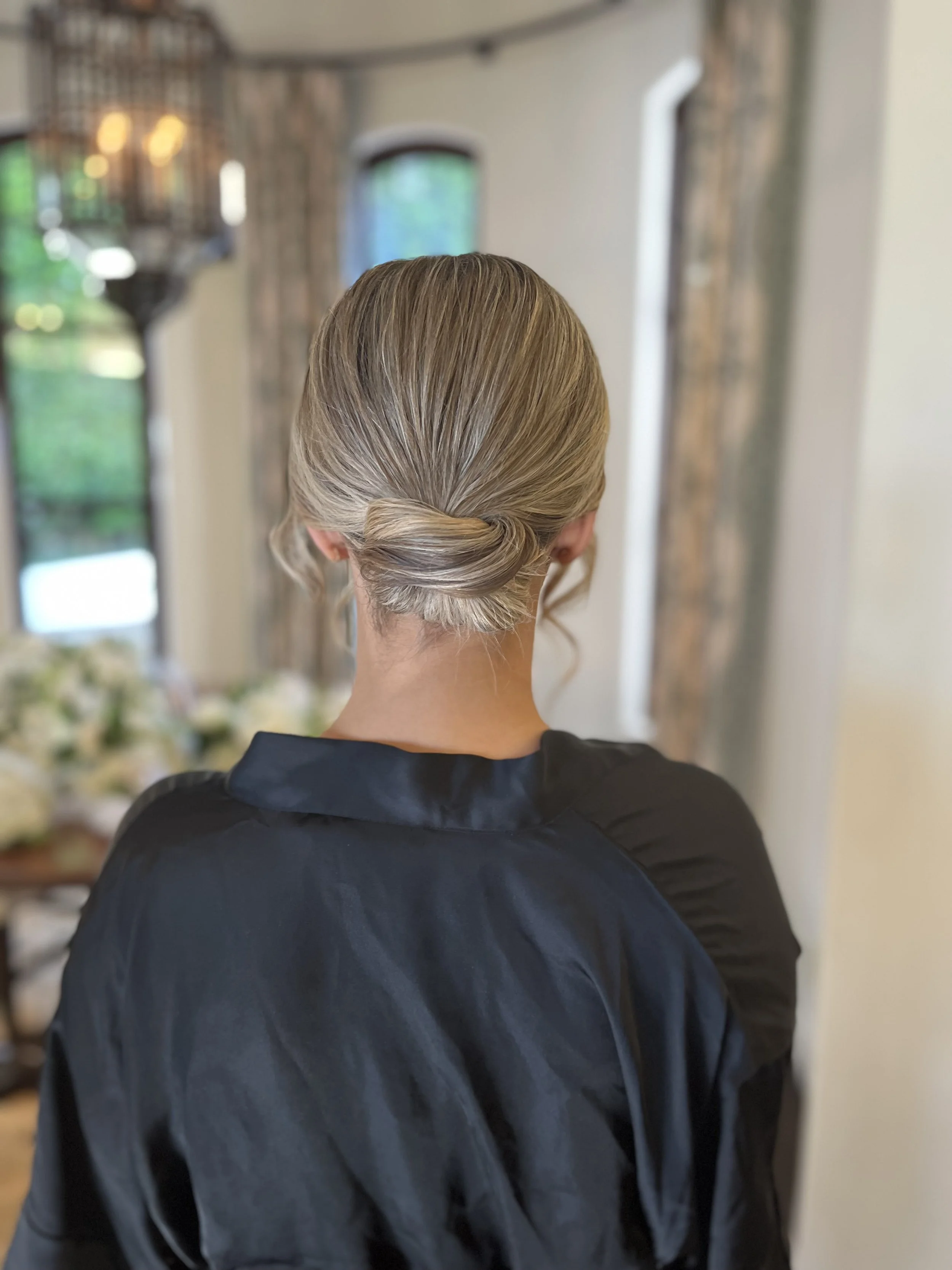 Back of a woman with blonde hair styled in a low bun, wearing a black jacket, in a room with blurred background of windows and curtains.