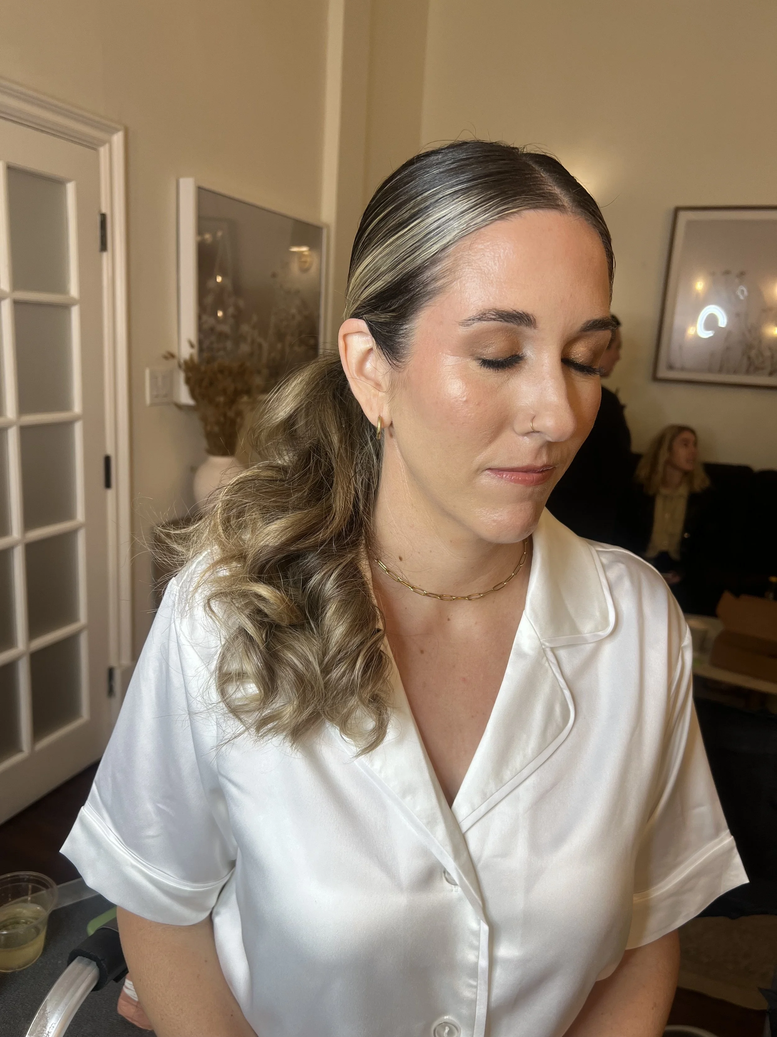 A woman with brown and blonde hair styled in waves, wearing a white satin blouse with a collar, gold hoop earrings, and a thin gold chain necklace, appears to be getting ready in a domestic setting.