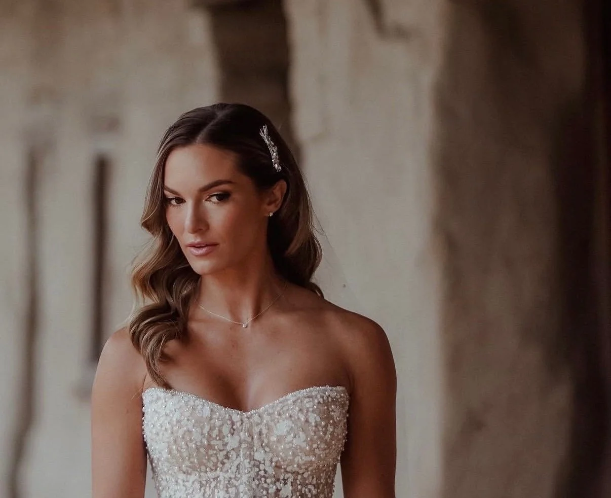 A woman in a strapless wedding dress with pearl embellishments, jewelry, and a hair accessory, standing indoors with a blurred stone wall background.