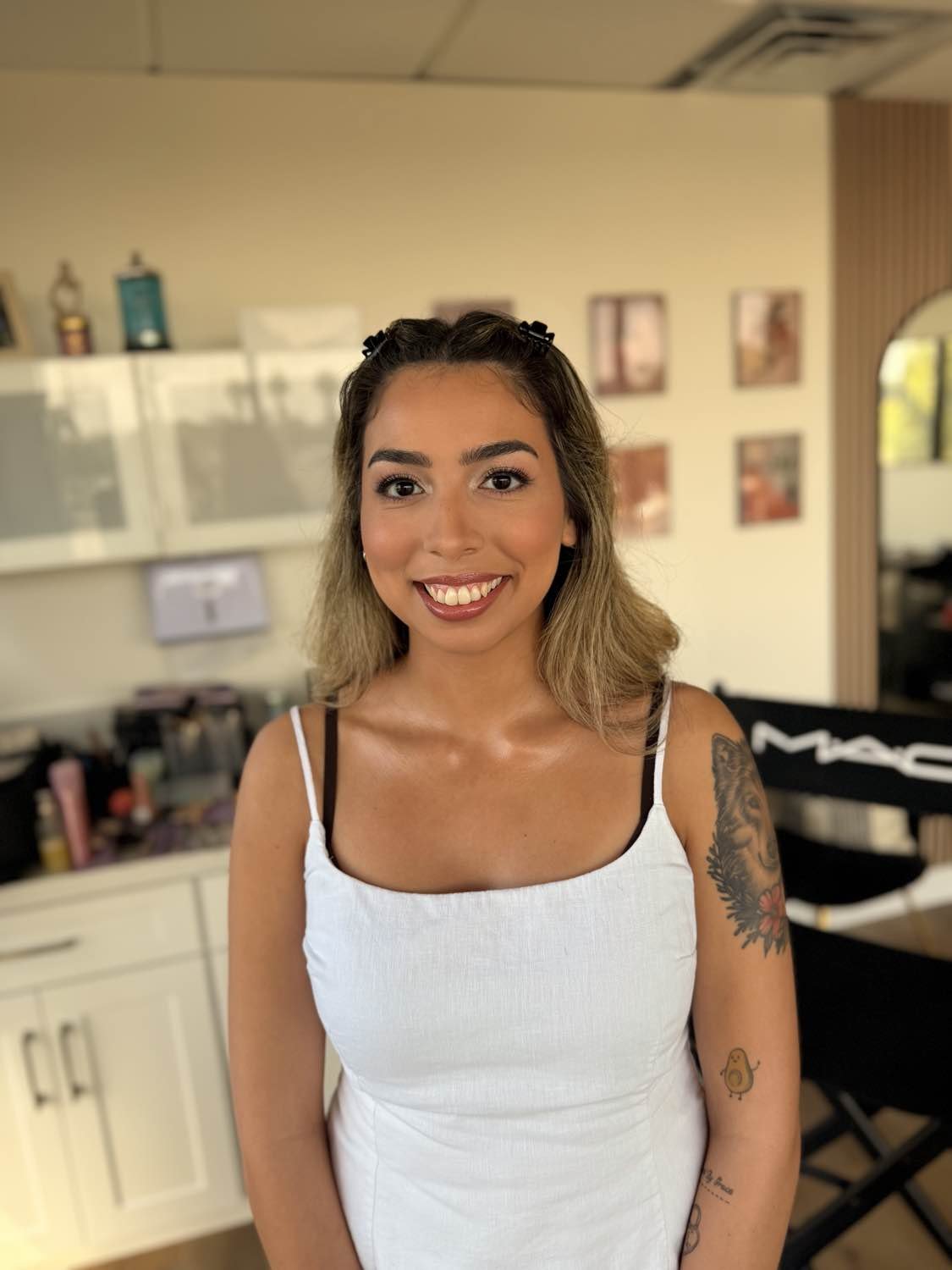 A woman with shoulder-length light brown hair, wearing a white tank top with black bra straps, smiling at the camera inside a room with framed pictures and shelves in the background.