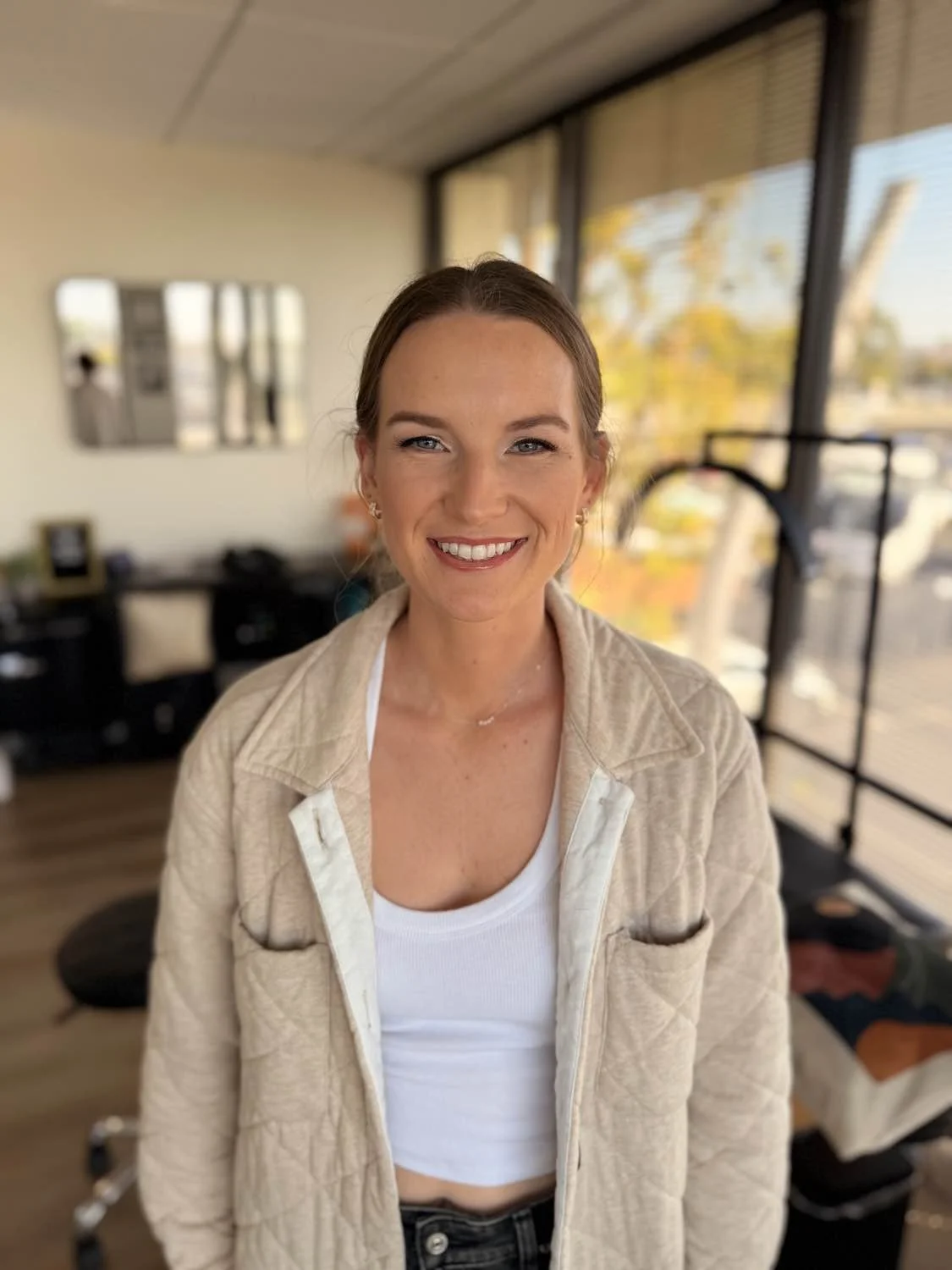 A woman with light brown hair, blue eyes, and a wide smile standing in a room with large windows and a city view, wearing a beige jacket over a white crop top.