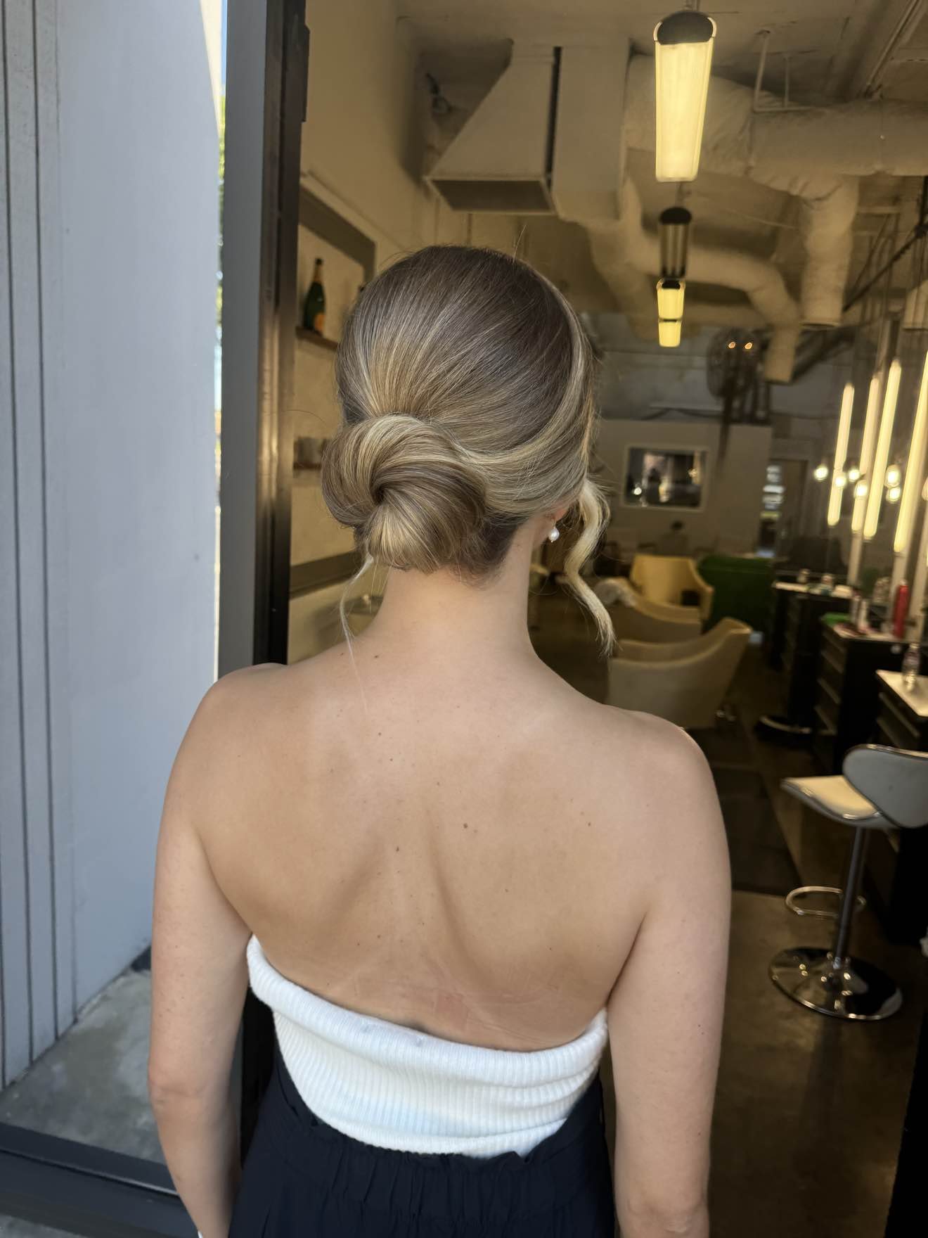 Back view of a woman with blonde hair in an elegant updo, standing in a modern indoor space, wearing a strapless white top and black pants.
