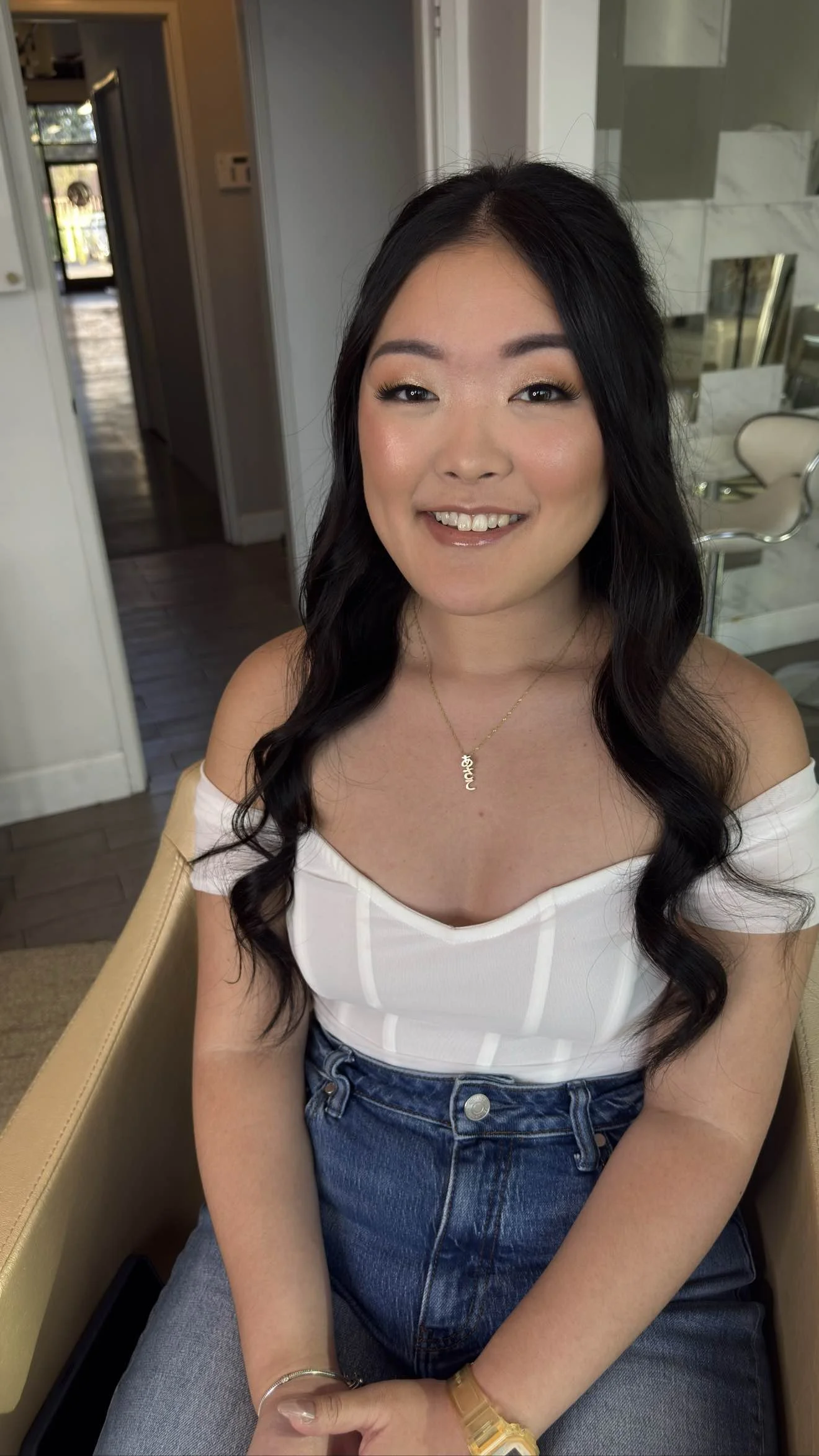 A woman with long dark wavy hair and light makeup sitting on a beige chair, smiling at the camera, wearing a white off-shoulder top, blue jeans, gold necklace, watch, and bracelet.